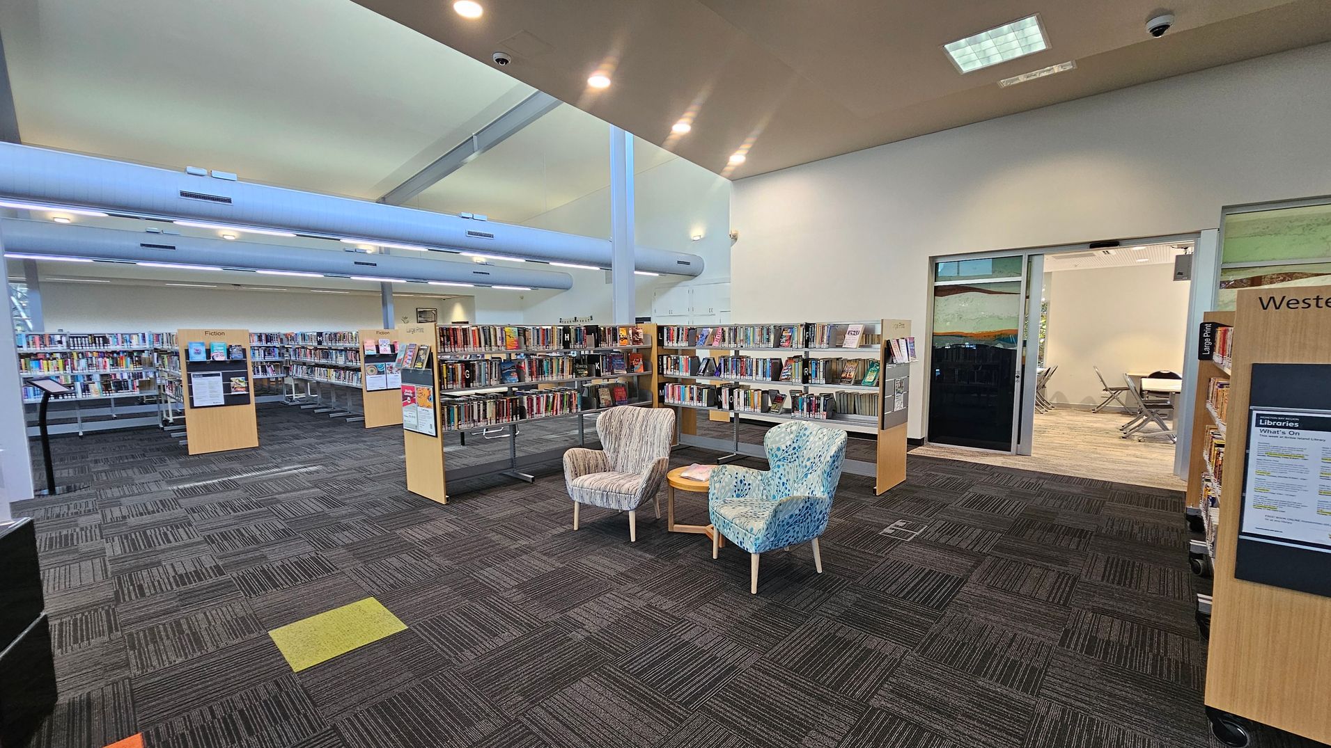 A library interior with bookshelves filled with books on the left and right. In the centre, two patterned armchairs are positioned around a small round table. The floor is covered with dark, patterned carpet tiles. In the background, a glass-walled room with tables and chairs is visible. The ceiling is high with recessed lighting and large, exposed ducts. A library interior with bookshelves filled with books on the left and right. In the centre, two patterned armchairs are positioned around a small round table. The floor is covered with dark, patterned carpet tiles. In the background, a glass-walled room with tables and chairs is visible. The ceiling is high with recessed lighting and large, exposed ducts.