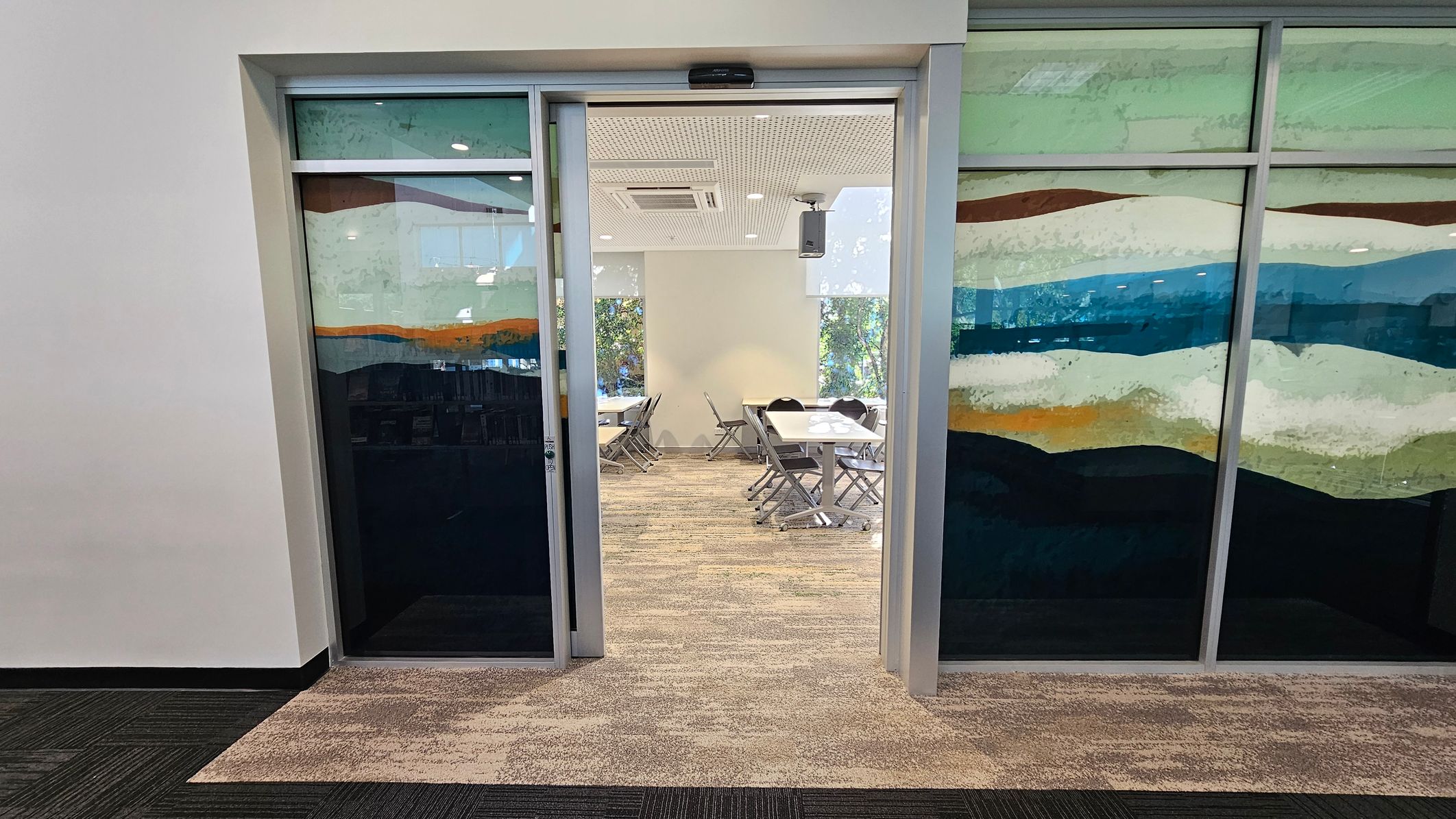 A room with glass sliding doors featuring a colourful abstract design. Inside, there are several tables and chairs arranged in a meeting or classroom style. The floor is covered with a patterned carpet, and the ceiling has recessed lighting and an air conditioning unit. The walls are light-coloured, and there are windows in the background allowing natural light to enter. A room with glass sliding doors featuring a colourful abstract design. Inside, there are several tables and chairs arranged in a meeting or classroom style. The floor is covered with a patterned carpet, and the ceiling has recessed lighting and an air conditioning unit. The walls are light-coloured, and there are windows in the background allowing natural light to enter.