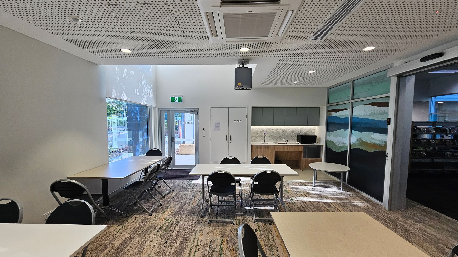 A room with a perforated ceiling featuring recessed lights and an air conditioning unit. On the left, large windows allow natural light in, illuminating tables and chairs. In the centre, a door marked "Store" is flanked by a kitchenette with cabinets, a sink, and a microwave. On the right, a wall with abstract art panels and a round table. The floor is carpeted with a patterned design. A room with a perforated ceiling featuring recessed lights and an air conditioning unit. On the left, large windows allow natural light in, illuminating tables and chairs. In the centre, a door marked "Store" is flanked by a kitchenette with cabinets, a sink, and a microwave. On the right, a wall with abstract art panels and a round table. The floor is carpeted with a patterned design.
