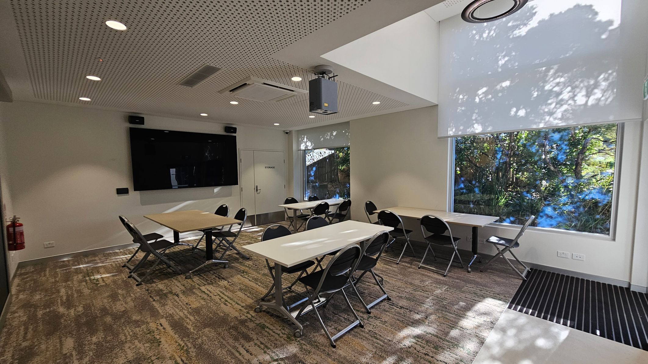 A modern conference room with a perforated ceiling featuring recessed lights and a projector. On the left, a large black screen is mounted on the wall above a door labelled "Storage". Several folding tables and chairs are arranged in the centre. On the right, large windows with roller blinds partially down allow natural light to filter through, casting shadows of trees outside. The floor is carpeted with a patterned design, and a fire extinguisher is mounted on the wall. A modern conference room with a perforated ceiling featuring recessed lights and a projector. On the left, a large black screen is mounted on the wall above a door labelled "Storage". Several folding tables and chairs are arranged in the centre. On the right, large windows with roller blinds partially down allow natural light to filter through, casting shadows of trees outside. The floor is carpeted with a patterned design, and a fire extinguisher is mounted on the wall.