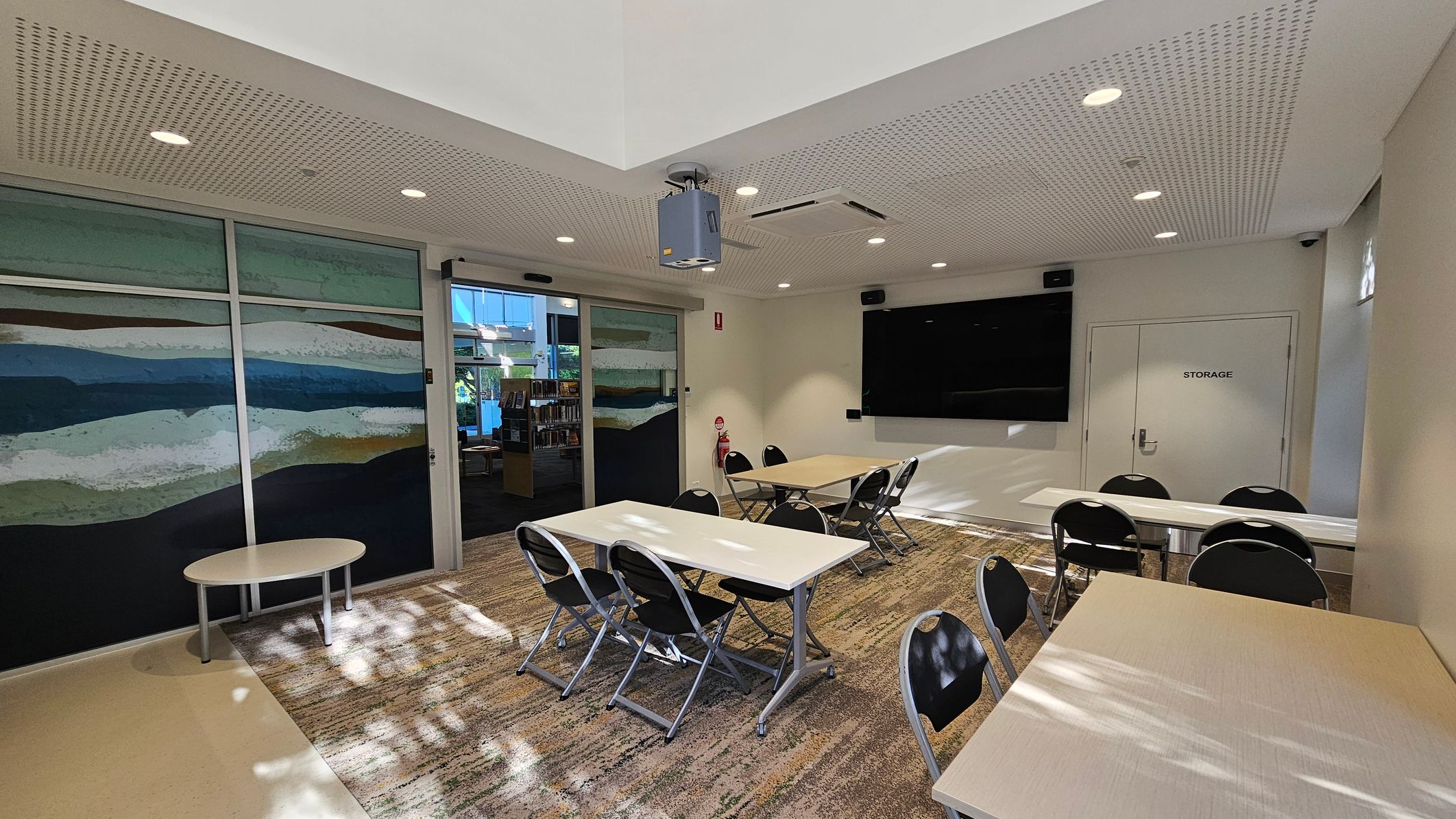 A room with a patterned carpet and a perforated ceiling with recessed lights. On the left, there is a large mural with abstract horizontal stripes in various colours. In the centre, several tables and chairs are arranged, with a projector mounted on the ceiling. On the right, a large black screen is mounted on the wall above a door labelled "STORAGE". A fire extinguisher is positioned near the door. In the background, a glass door leads to another area with bookshelves. A room with a patterned carpet and a perforated ceiling with recessed lights. On the left, there is a large mural with abstract horizontal stripes in various colours. In the centre, several tables and chairs are arranged, with a projector mounted on the ceiling. On the right, a large black screen is mounted on the wall above a door labelled "STORAGE". A fire extinguisher is positioned near the door. In the background, a glass door leads to another area with bookshelves.