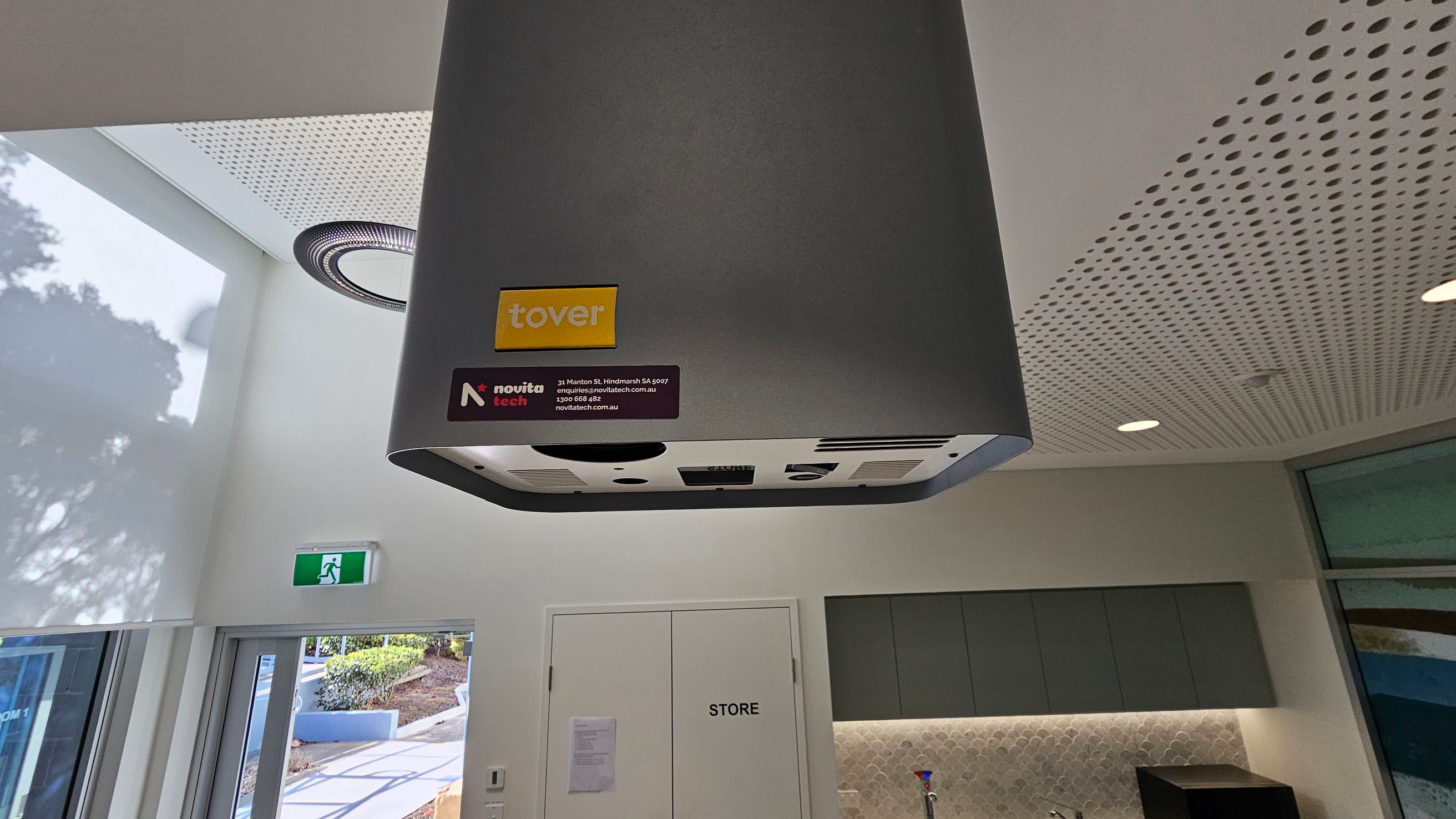 A grey ceiling-mounted device with "tover" and "novita tech" labels is in the foreground. The ceiling has a perforated design with recessed lighting. Below, a white door marked "STORE" is flanked by grey cabinets and a countertop with a sink. On the left, a glass door leads outside, where a path and greenery are visible. An emergency exit sign is above the door. A grey ceiling-mounted device with "tover" and "novita tech" labels is in the foreground. The ceiling has a perforated design with recessed lighting. Below, a white door marked "STORE" is flanked by grey cabinets and a countertop with a sink. On the left, a glass door leads outside, where a path and greenery are visible. An emergency exit sign is above the door.
