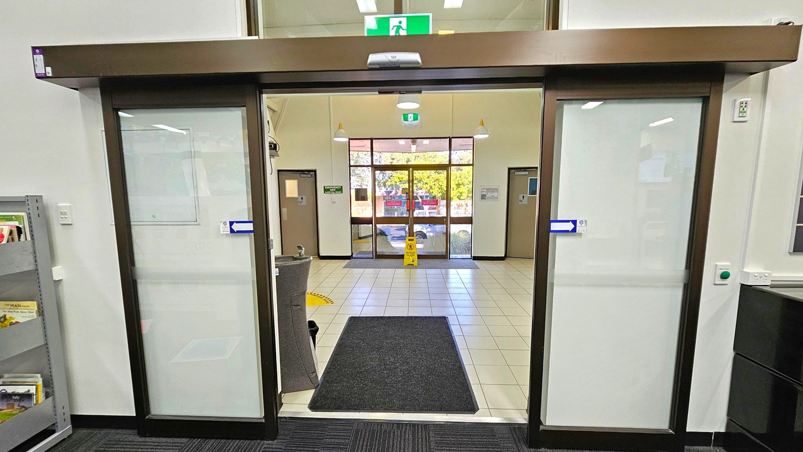 Automatic sliding glass doors with a dark frame are in the foreground, leading to a tiled entrance area. A black mat is on the floor just inside the doors. Beyond, a larger set of glass doors opens to the outside, with a yellow caution sign on the floor. On the left, a grey shelving unit holds books, and on the right, a wall with a green button and power outlet. The ceiling has recessed lighting and a green emergency exit sign above the doors. Automatic sliding glass doors with a dark frame are in the foreground, leading to a tiled entrance area. A black mat is on the floor just inside the doors. Beyond, a larger set of glass doors opens to the outside, with a yellow caution sign on the floor. On the left, a grey shelving unit holds books, and on the right, a wall with a green button and power outlet. The ceiling has recessed lighting and a green emergency exit sign above the doors.