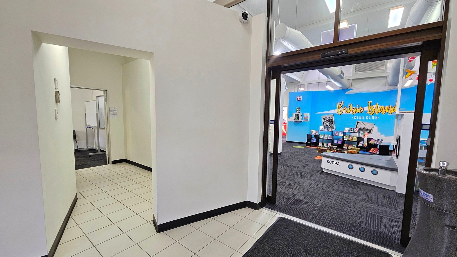 A hallway with white tiled flooring and white walls leads to a room on the left with a whiteboard visible inside. On the right, a doorway opens into a room with dark carpeted flooring. The back wall is bright blue with the words "Bribie Island Kids Club" in yellow. Below the sign, there are shelves displaying books. A play kitchen labelled "Koopa" is in the foreground. The ceiling has exposed ducts and lights, and a water fountain is visible on the right. A hallway with white tiled flooring and white walls leads to a room on the left with a whiteboard visible inside. On the right, a doorway opens into a room with dark carpeted flooring. The back wall is bright blue with the words "Bribie Island Kids Club" in yellow. Below the sign, there are shelves displaying books. A play kitchen labelled "Koopa" is in the foreground. The ceiling has exposed ducts and lights, and a water fountain is visible on the right.
