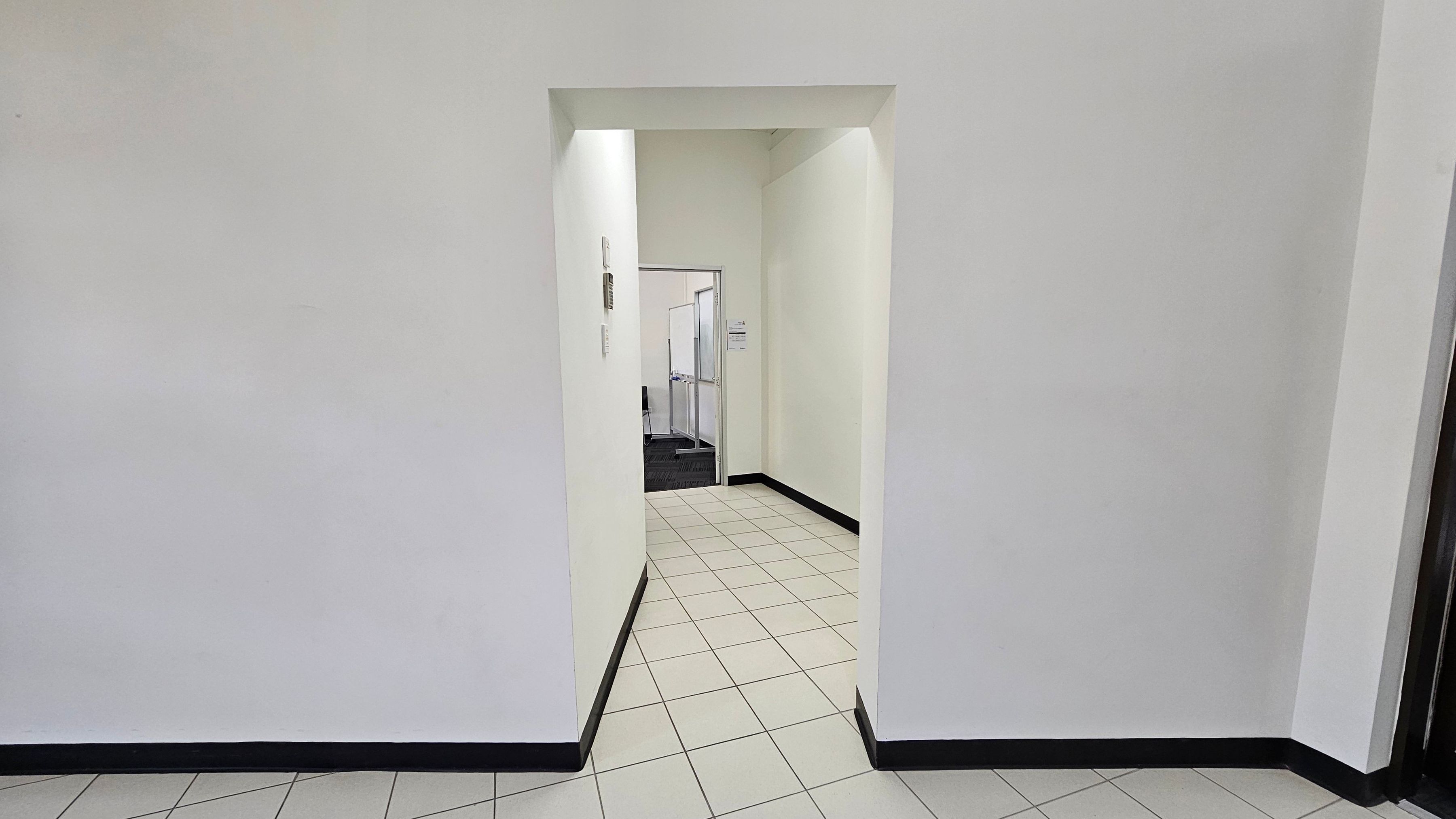 A hallway with white walls and a tiled floor leading to an open doorway. Beyond the doorway, there is a room with a darker carpeted floor. The walls have a slight yellow tint, and there are some fixtures on the left wall near the doorway. The ceiling is plain and white, with no visible features. A hallway with white walls and a tiled floor leading to an open doorway. Beyond the doorway, there is a room with a darker carpeted floor. The walls have a slight yellow tint, and there are some fixtures on the left wall near the doorway. The ceiling is plain and white, with no visible features.