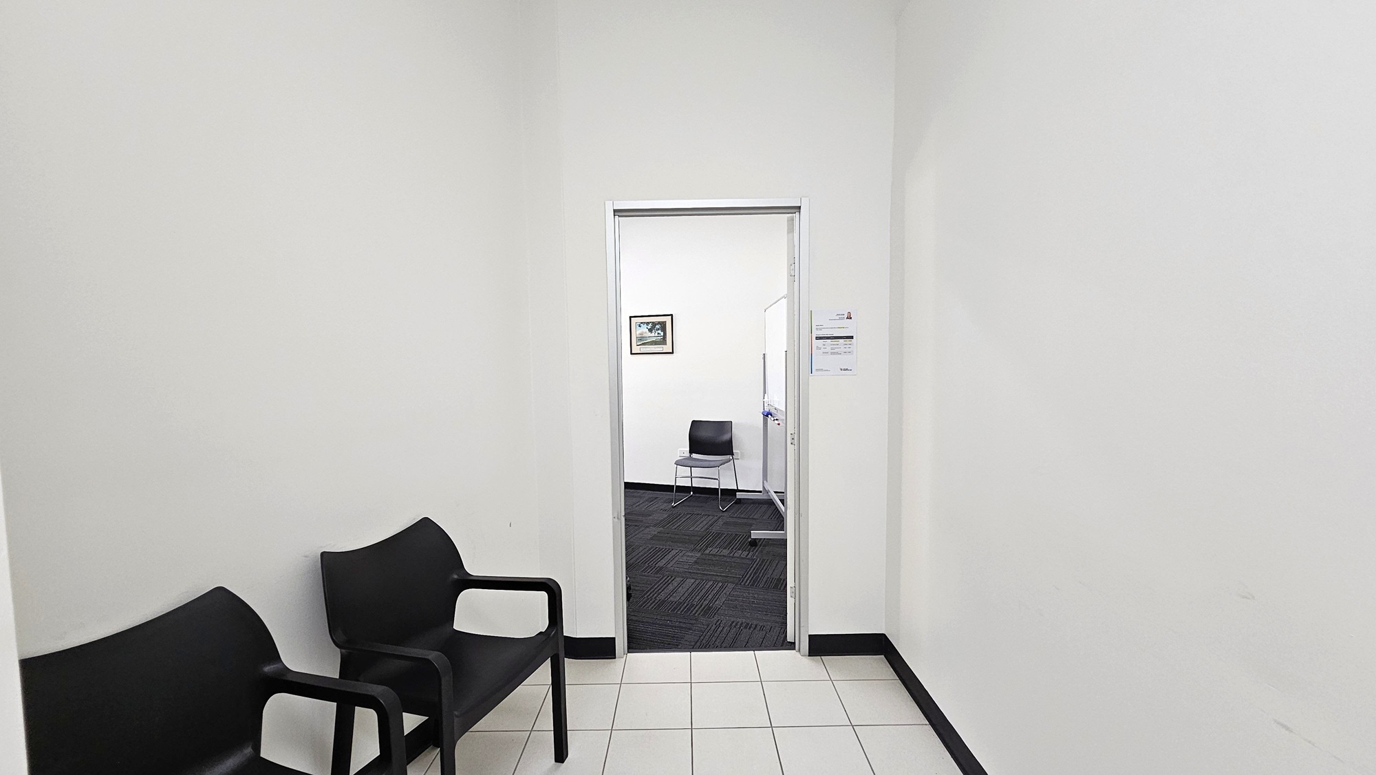A hallway with white walls and a tiled floor. On the left, there are two black chairs. Ahead, an open doorway leads to another room with a dark carpeted floor. Inside, a single black chair is visible against the far wall. A framed picture hangs on the wall to the left, and a whiteboard is partially visible on the right. A small sign is posted on the wall near the doorway. A hallway with white walls and a tiled floor. On the left, there are two black chairs. Ahead, an open doorway leads to another room with a dark carpeted floor. Inside, a single black chair is visible against the far wall. A framed picture hangs on the wall to the left, and a whiteboard is partially visible on the right. A small sign is posted on the wall near the doorway.