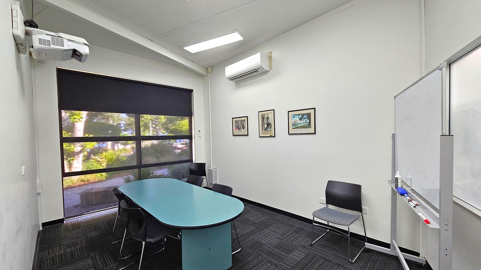 A small meeting room with a black carpeted floor and a white ceiling. On the left, a window with a black roller blind partially open, revealing greenery outside. Below the window is a blue oval table surrounded by black chairs. On the right wall, three framed pictures are displayed. An air conditioning unit is mounted above them. A whiteboard with markers is positioned on the far right. A projector is mounted on the ceiling. A small meeting room with a black carpeted floor and a white ceiling. On the left, a window with a black roller blind partially open, revealing greenery outside. Below the window is a blue oval table surrounded by black chairs. On the right wall, three framed pictures are displayed. An air conditioning unit is mounted above them. A whiteboard with markers is positioned on the far right. A projector is mounted on the ceiling.