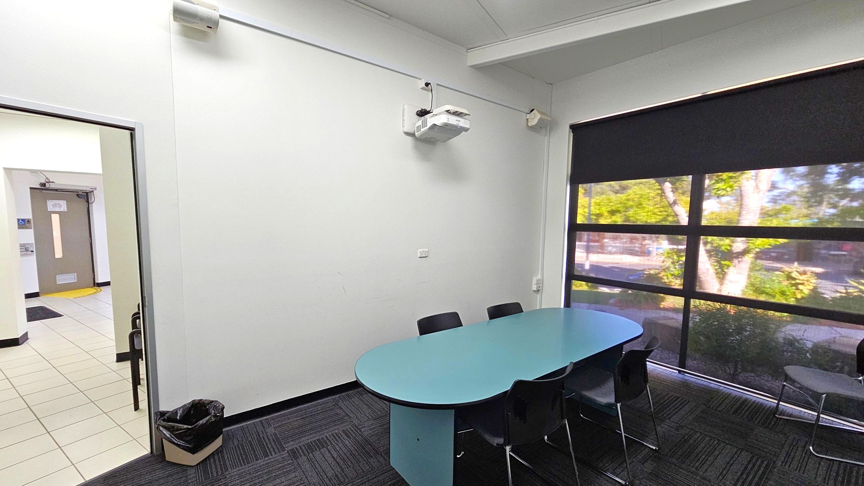 A small meeting room with a blue oval table surrounded by black chairs on the right. A large window with a black blind partially drawn reveals trees outside. On the left, an open door leads to a tiled hallway with a brown door visible. A black bin is on the floor near the door. A white projector is mounted on the wall above the table. The floor is carpeted with a dark pattern, and the ceiling is white with visible beams. A small meeting room with a blue oval table surrounded by black chairs on the right. A large window with a black blind partially drawn reveals trees outside. On the left, an open door leads to a tiled hallway with a brown door visible. A black bin is on the floor near the door. A white projector is mounted on the wall above the table. The floor is carpeted with a dark pattern, and the ceiling is white with visible beams.
