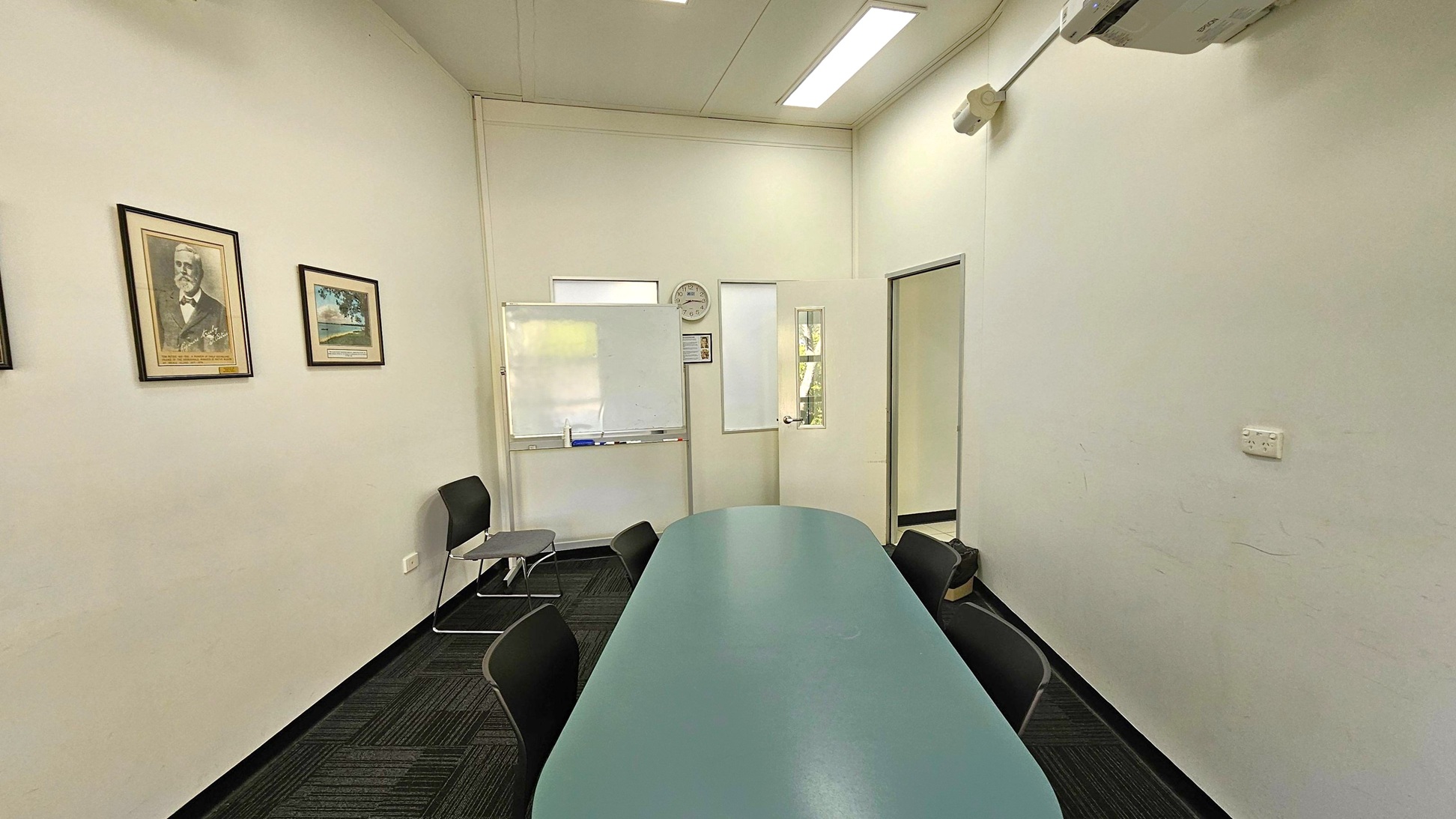 A small meeting room with a long oval table in the centre surrounded by black chairs. On the left wall, there are three framed pictures. The back wall features a whiteboard, a clock, and a door with a window. The right wall has an air conditioning unit mounted near the ceiling and a power outlet. The floor is carpeted in a dark pattern, and the ceiling has recessed lighting. A small meeting room with a long oval table in the centre surrounded by black chairs. On the left wall, there are three framed pictures. The back wall features a whiteboard, a clock, and a door with a window. The right wall has an air conditioning unit mounted near the ceiling and a power outlet. The floor is carpeted in a dark pattern, and the ceiling has recessed lighting.