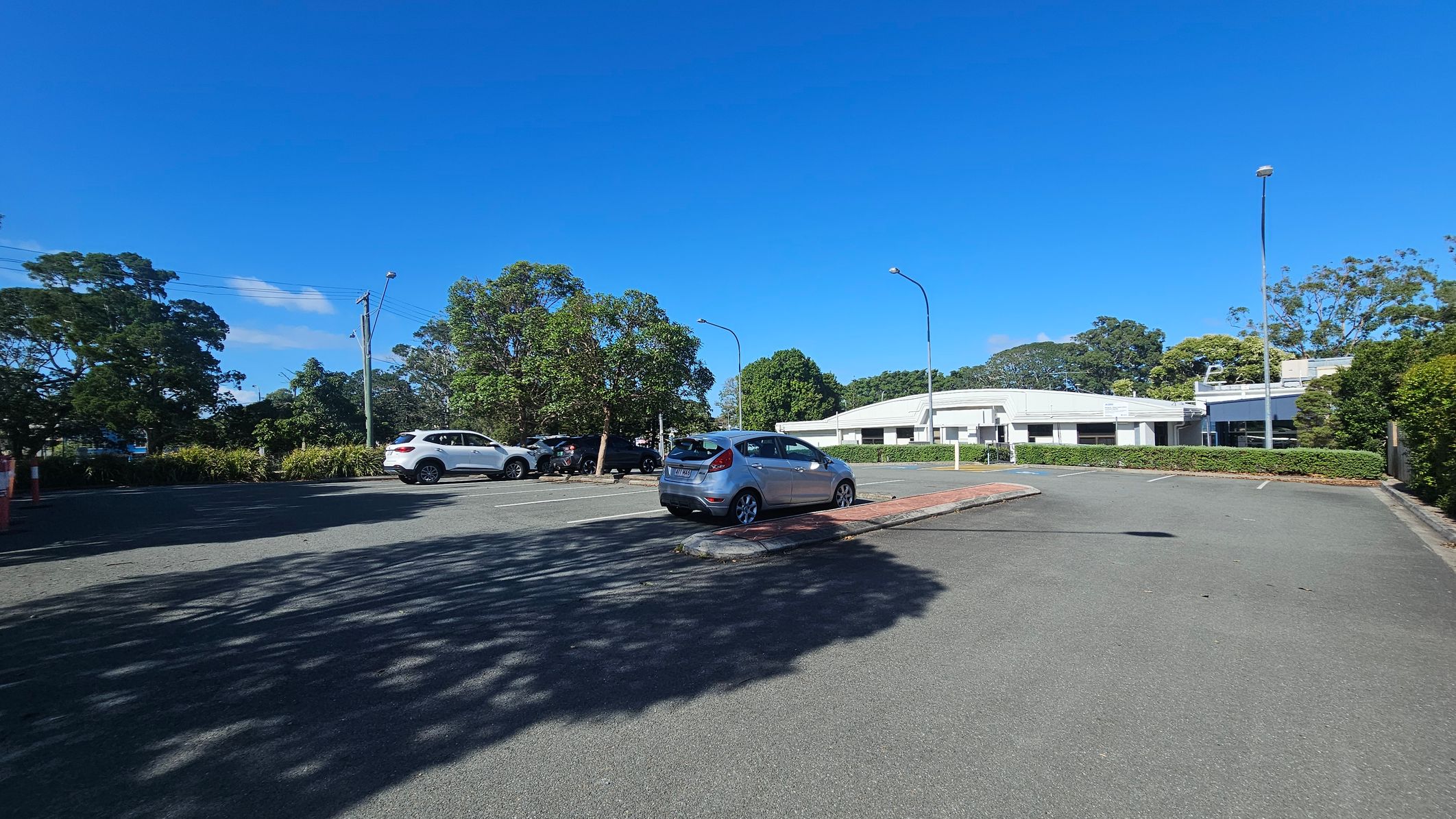 A car park with a few vehicles, including a silver car in the centre. On the left, there are trees and a white SUV. In the background, a white building is partially visible, surrounded by more trees and greenery. The ground is paved, and there are streetlights along the perimeter. Shadows of trees are cast on the pavement. A car park with a few vehicles, including a silver car in the centre. On the left, there are trees and a white SUV. In the background, a white building is partially visible, surrounded by more trees and greenery. The ground is paved, and there are streetlights along the perimeter. Shadows of trees are cast on the pavement.
