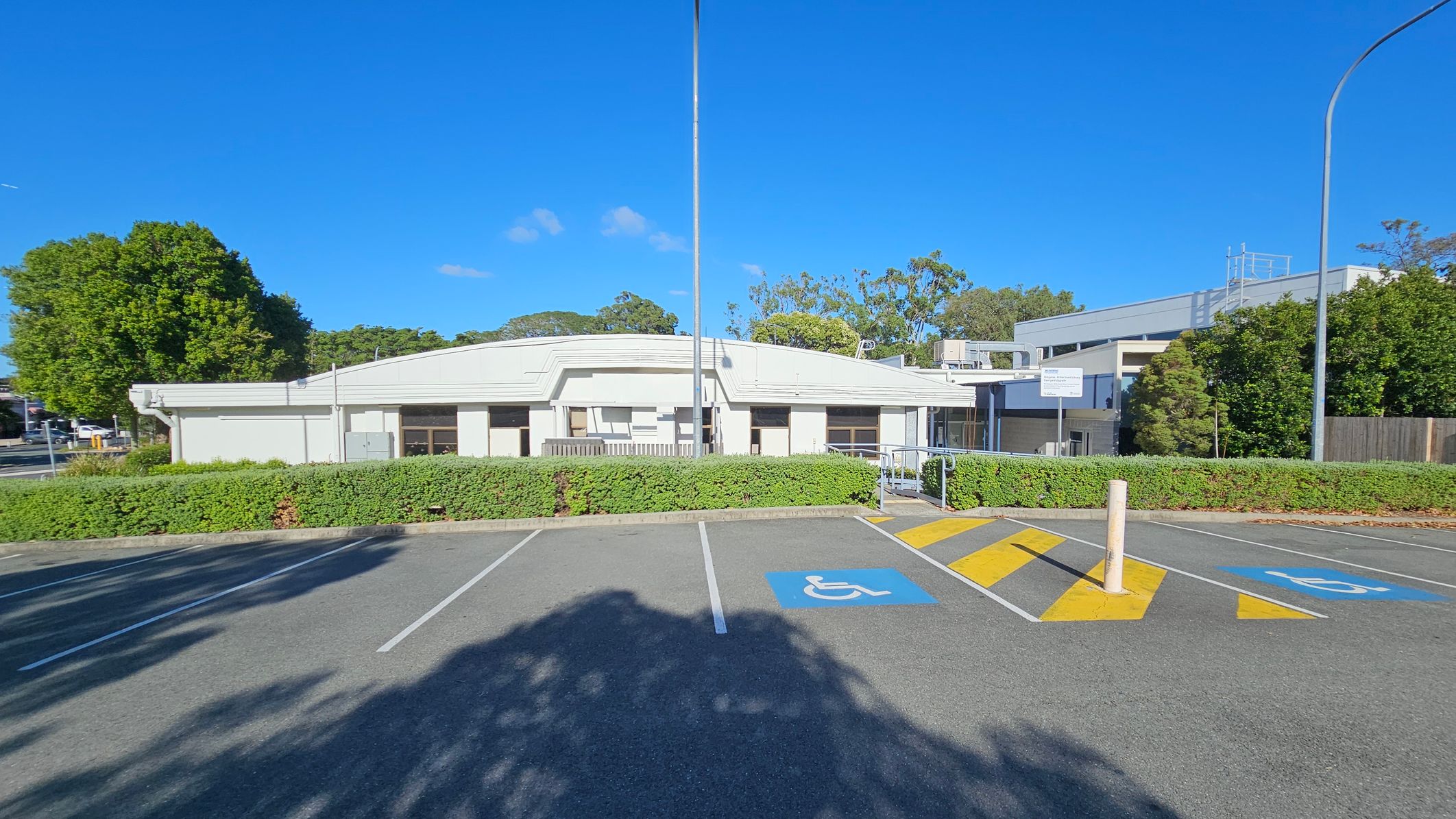 A single-storey white building with a flat roof is in the centre, surrounded by greenery. In the foreground, there is a car park with marked disabled parking spaces and a yellow-striped pedestrian crossing. Tall streetlights are visible on either side. Trees and bushes are in the background, with a clear blue sky above. A single-storey white building with a flat roof is in the centre, surrounded by greenery. In the foreground, there is a car park with marked disabled parking spaces and a yellow-striped pedestrian crossing. Tall streetlights are visible on either side. Trees and bushes are in the background, with a clear blue sky above.