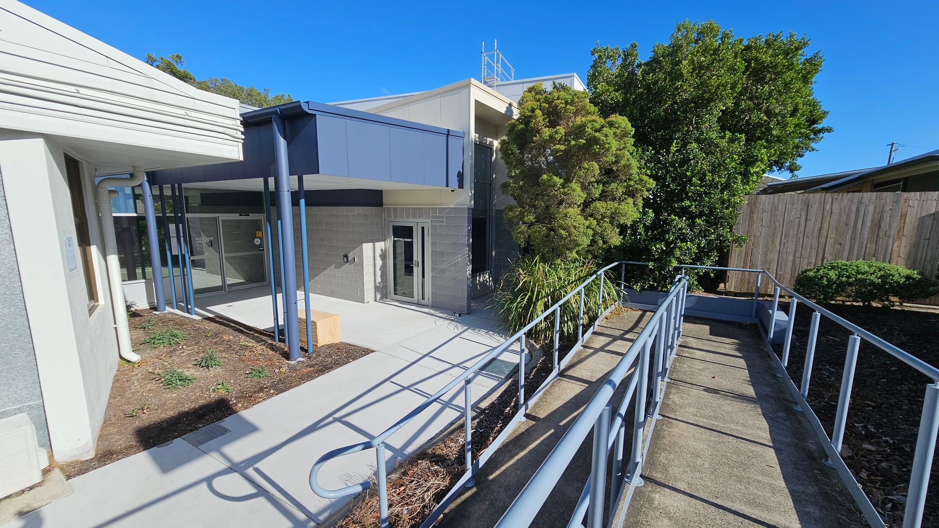 A building entrance with a ramp leading up to it. On the left, there is a white and grey wall with a window and a downpipe. The middle features a glass door with a sign, supported by blue pillars and a small garden area with plants. On the right, there is a wooden fence and dense greenery, including a large bush and smaller shrubs. The ramp has metal railings and is bordered by a narrow garden bed. The ground is paved with concrete. A building entrance with a ramp leading up to it. On the left, there is a white and grey wall with a window and a downpipe. The middle features a glass door with a sign, supported by blue pillars and a small garden area with plants. On the right, there is a wooden fence and dense greenery, including a large bush and smaller shrubs. The ramp has metal railings and is bordered by a narrow garden bed. The ground is paved with concrete.