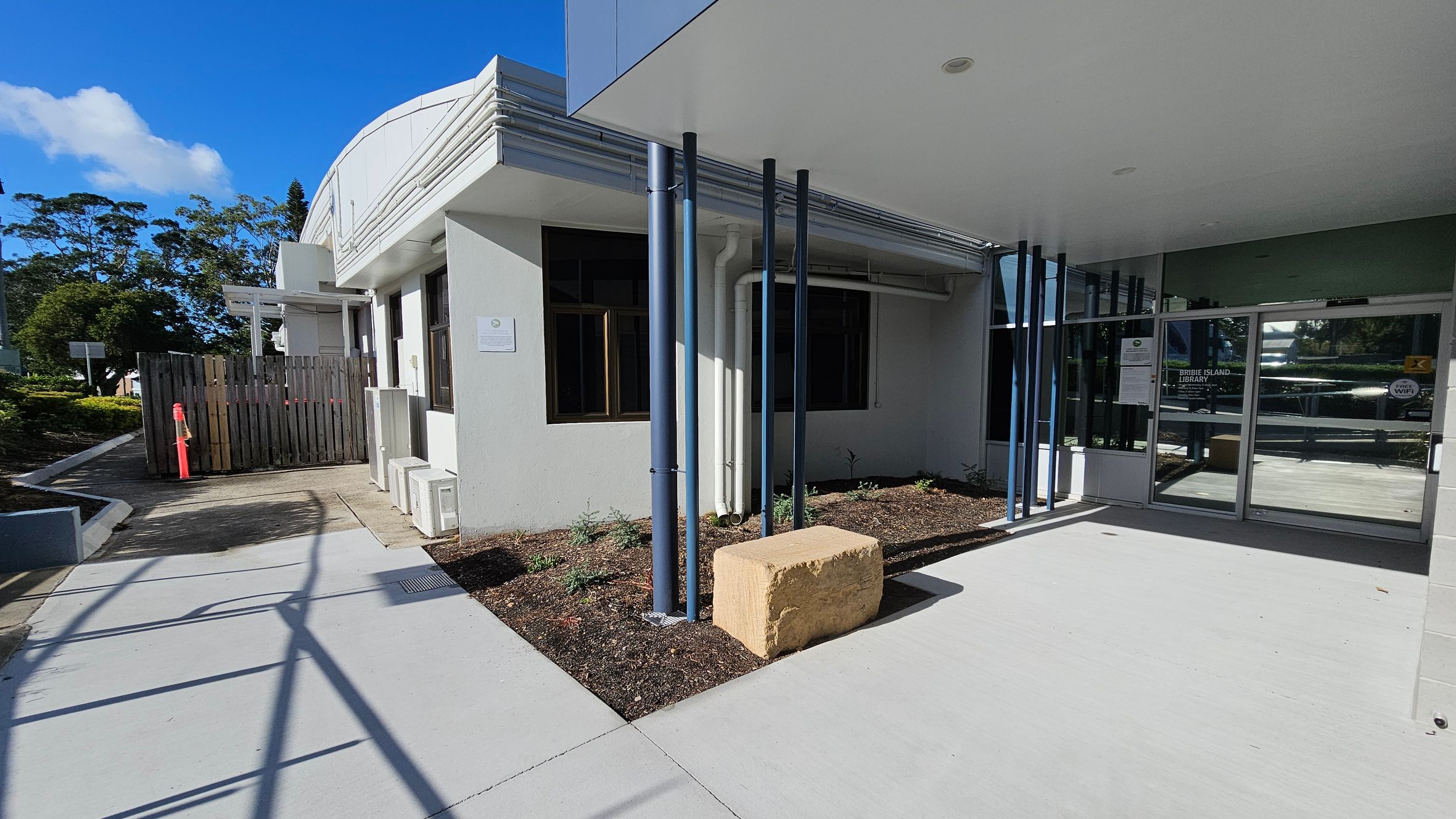 A modern building entrance with a glass door on the right, featuring signage. Blue vertical pipes are positioned near the entrance, with a large rectangular stone block beside them. The left side has a wooden fence and a pathway leading away from the building. The ground is concrete, and there are small plants in a mulched area near the building. The ceiling is flat and white, with a recessed light. Trees and greenery are visible in the background. A modern building entrance with a glass door on the right, featuring signage. Blue vertical pipes are positioned near the entrance, with a large rectangular stone block beside them. The left side has a wooden fence and a pathway leading away from the building. The ground is concrete, and there are small plants in a mulched area near the building. The ceiling is flat and white, with a recessed light. Trees and greenery are visible in the background.