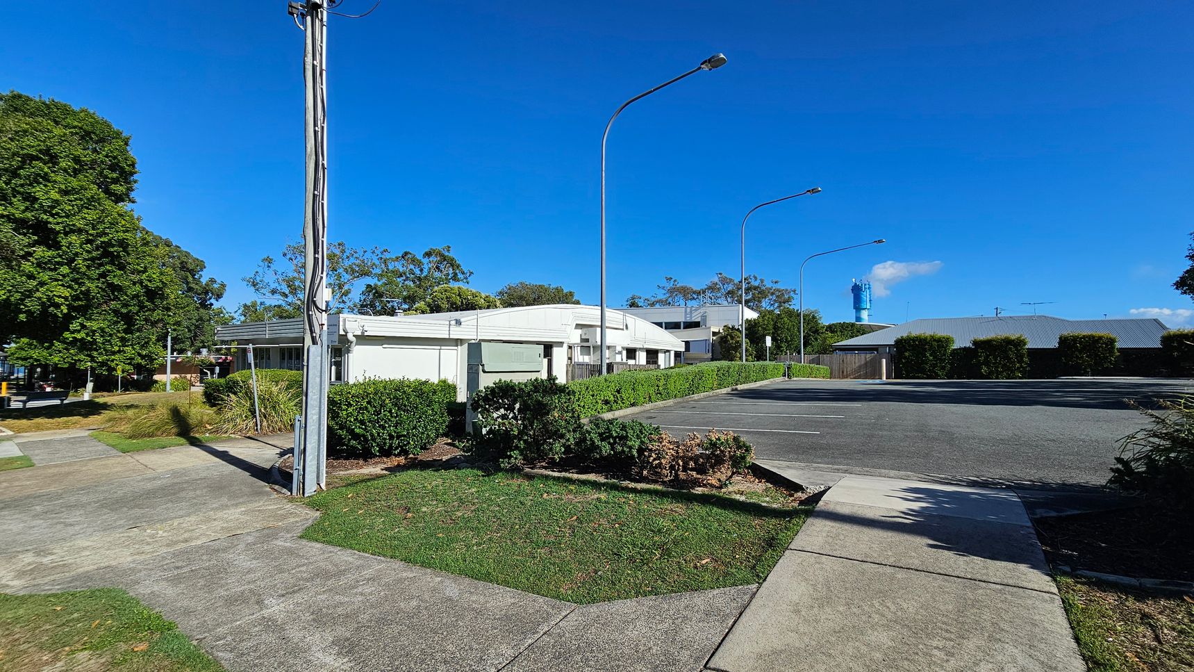 A street scene with a white building in the centre, surrounded by green hedges and trees. On the left, a tall utility pole stands near a grassy area and a concrete footpath. The right side features a paved parking area with several streetlights. In the background, a blue structure is visible, partially obscured by trees and buildings. The sky is clear and blue. A street scene with a white building in the centre, surrounded by green hedges and trees. On the left, a tall utility pole stands near a grassy area and a concrete footpath. The right side features a paved parking area with several streetlights. In the background, a blue structure is visible, partially obscured by trees and buildings. The sky is clear and blue.