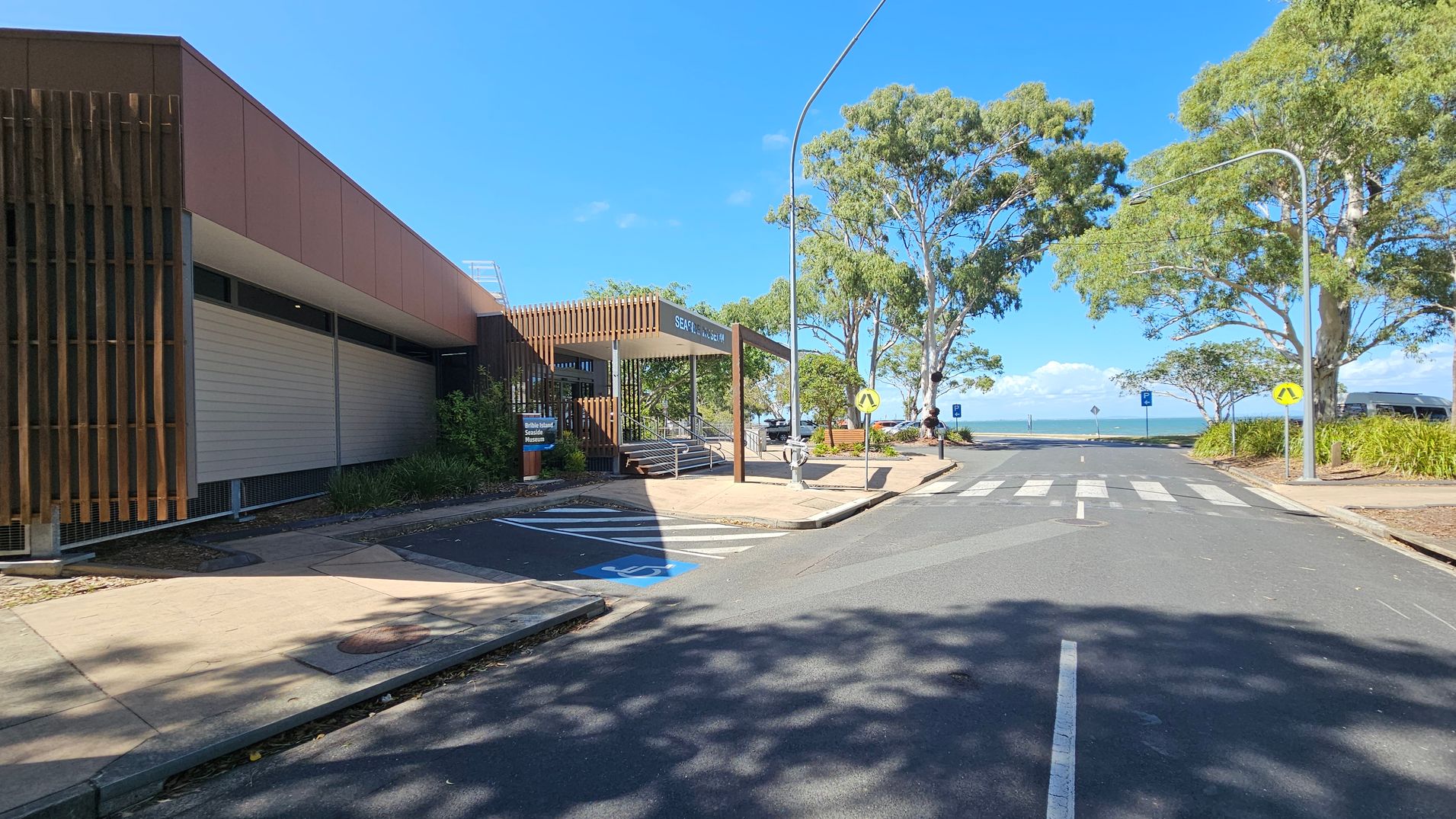 A street scene with a building on the left featuring vertical wooden slats and a sign reading "Seafood Market". A pedestrian crossing leads towards the sea in the background. Trees line the right side of the street, providing shade. A blue disabled parking space is visible in the foreground. The sky is clear and blue, and there are street lamps along the road.