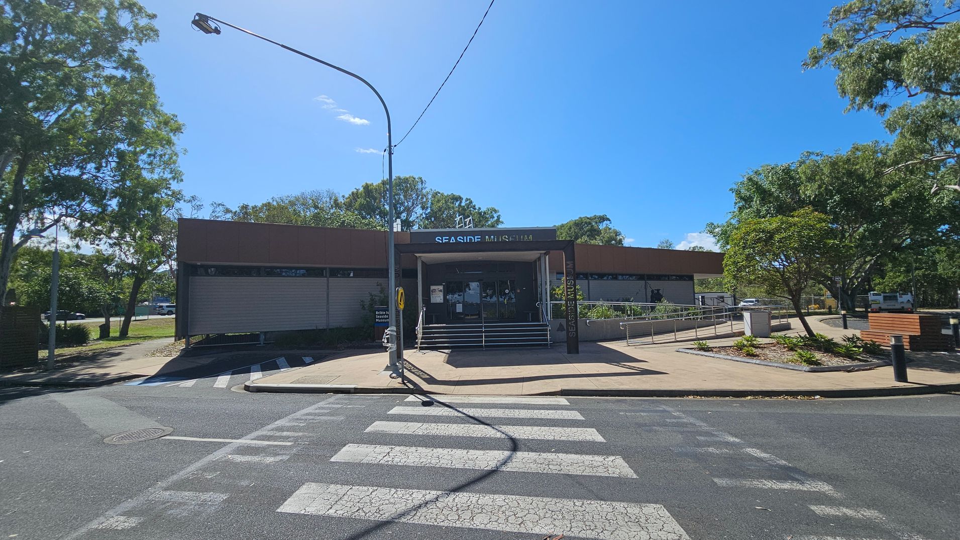 A single-storey building with a sign reading "Seaside Museum" above the entrance. The entrance has steps and a ramp, with metal railings. A zebra crossing leads to the entrance from the foreground. Trees and greenery are on both sides, with a streetlamp on the left. The pavement is light-coloured, and the road is dark grey. A bench and a small landscaped area with plants are on the right.