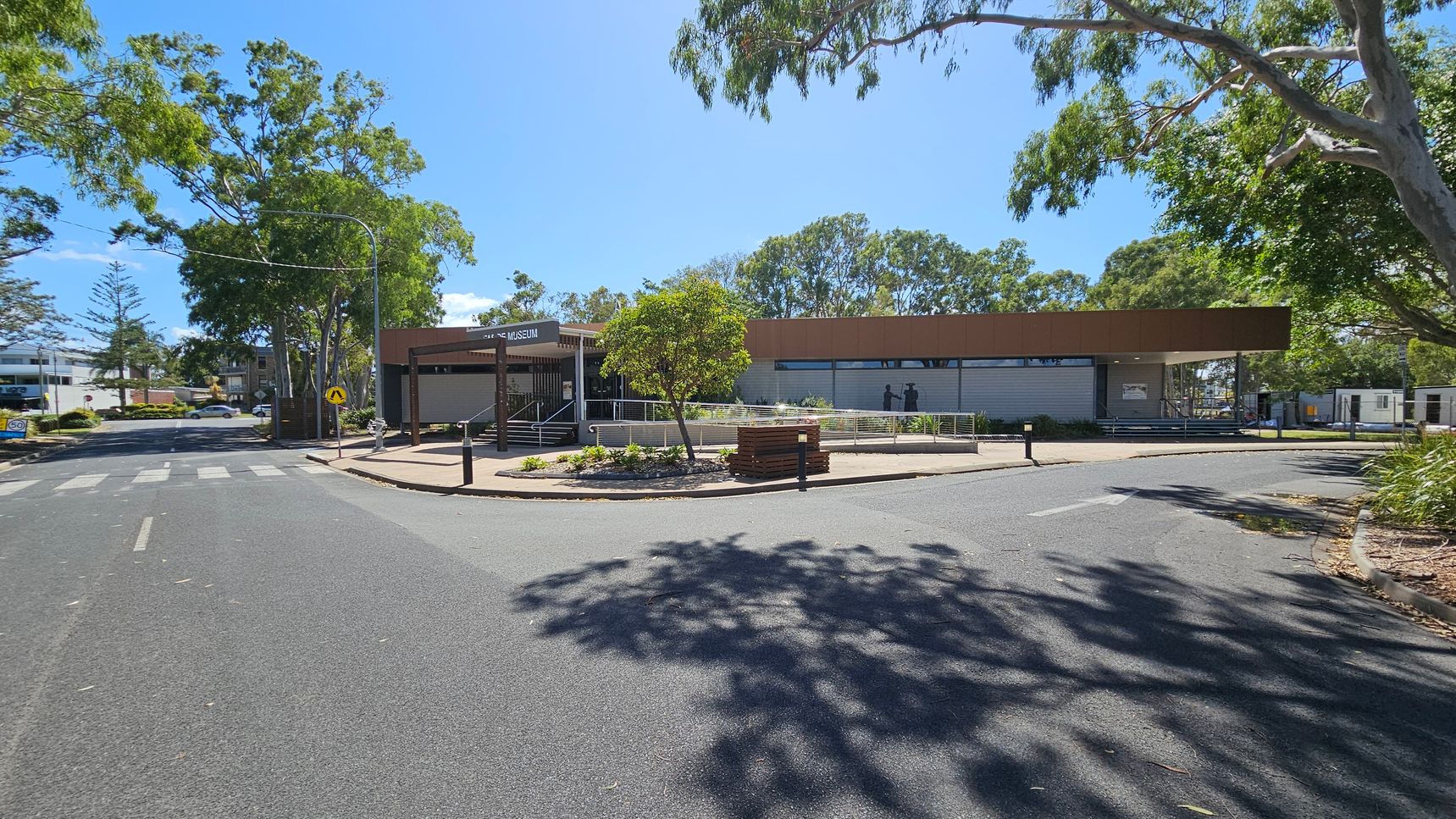A street scene with a museum building in the centre, featuring a brown roof and a sign. A pedestrian crossing is visible on the road in the foreground. Trees with green foliage are on both sides, casting shadows on the pavement. To the left, there are residential buildings and a speed limit sign. The area is landscaped with small plants and a bench near the museum entrance. The sky is clear and blue.