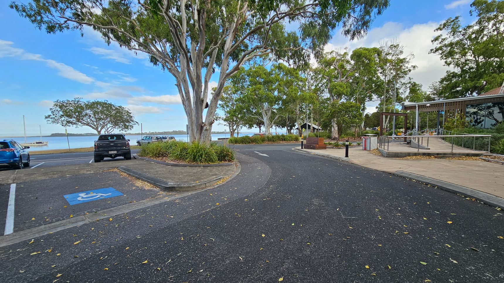 A paved road curves through a park-like area. On the left, there are parked cars, including a blue car and a black vehicle in a disabled parking space. Trees with green foliage line the area, providing shade. On the right, a modern building with large glass windows and a ramp is visible. The background features a body of water with a boat and distant land. The ground is scattered with fallen leaves.