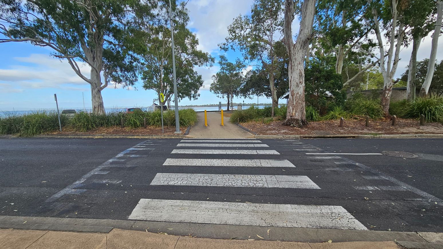 A zebra crossing leads to a path flanked by two yellow bollards. Tall trees with thick trunks and green foliage line both sides of the path. The ground is covered with grass and mulch. In the background, a body of water is visible, bordered by more trees and vegetation. The sky is partly cloudy. The road surface is dark with visible wear, and the pavement is light brown.