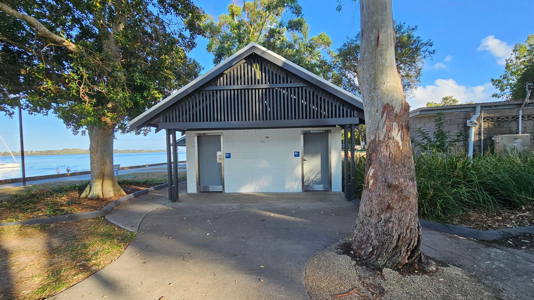 A small public restroom building with a gabled roof and two doors, each marked with a blue sign. The structure is surrounded by trees, with a large tree trunk in the foreground on the right. To the left, a path leads towards a waterfront area with a couple of people near the water and a boat in the distance. The ground is a mix of concrete and natural earth, and there is a grassy area with plants on the right side.