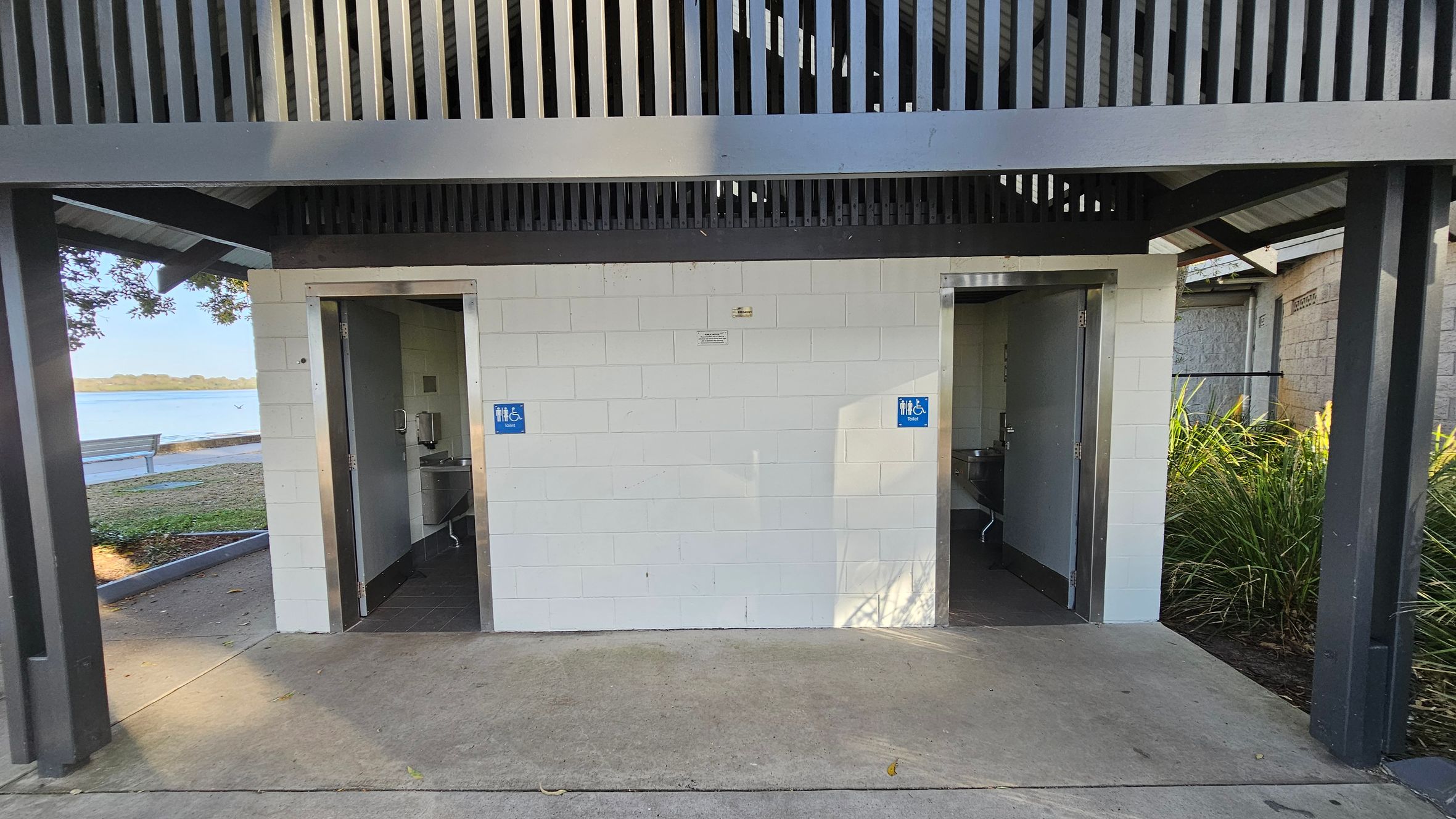 Two public toilet entrances with open doors are visible, each marked with blue signs indicating accessibility. The structure is made of white bricks with a grey metal roof featuring vertical slats. On the left, a glimpse of a bench and water body is visible through the trees. On the right, there are green plants. The floor is concrete, and the ceiling is not visible.