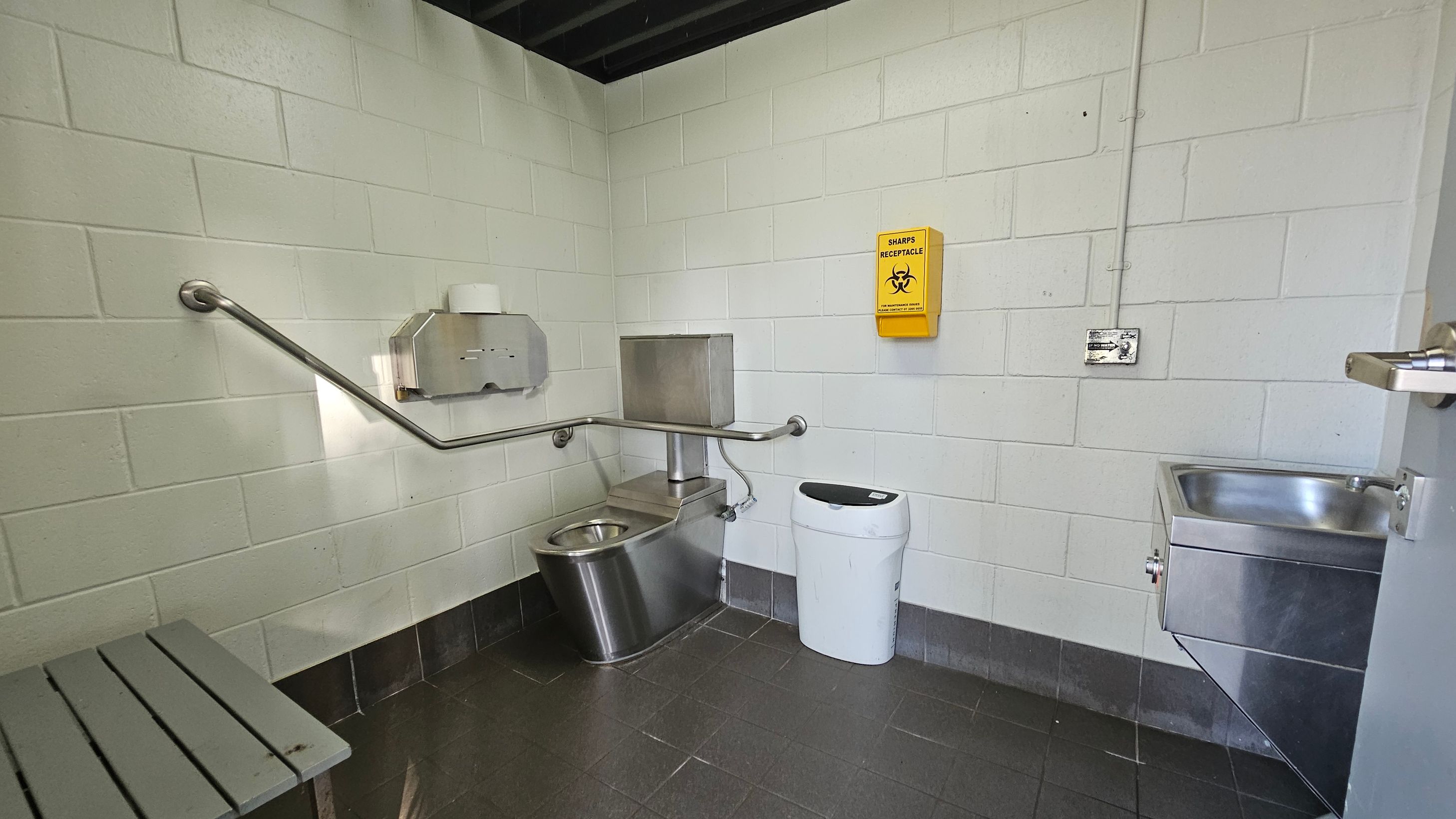 A small, utilitarian bathroom with white tiled walls and a dark tiled floor. On the left, a metal bench is visible. In the centre, there is a stainless steel toilet with a metal toilet paper dispenser above it and a grab bar along the wall. A yellow sharps receptacle is mounted on the wall. To the right, a small stainless steel sink is attached to the wall, and a white bin is placed nearby. The ceiling is dark and industrial-looking.