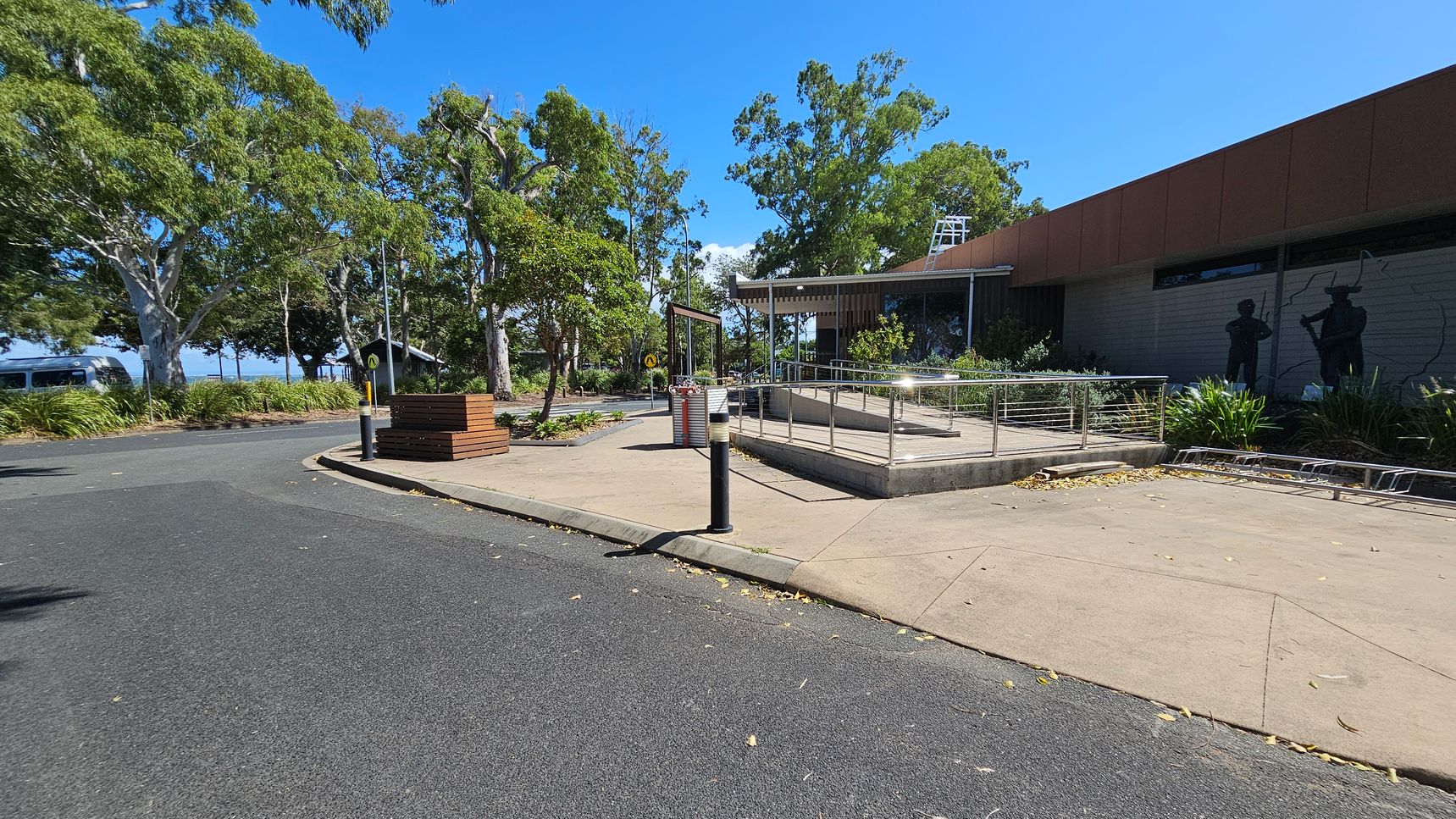 A paved area with a ramp and metal railing is on the right, leading to a building with a red roof. Silhouettes of figures are visible on the building's wall. Trees with green foliage are scattered throughout the background. On the left, a curved road is bordered by grass and more trees. A bench and a small shelter are visible near the road. The ground is covered with a mix of asphalt and concrete, and a few bollards are present.