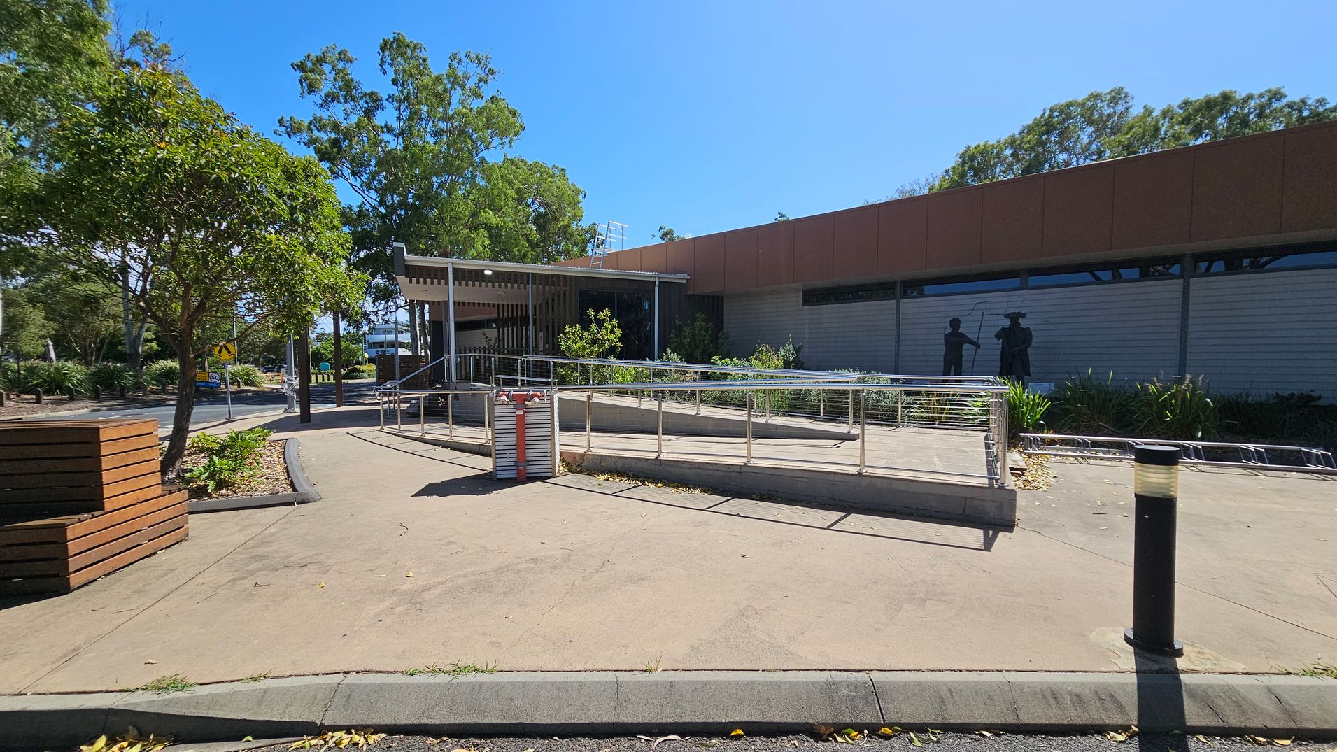 A paved area with a metal railing ramp in the centre. To the right, a building with a red-brown facade and a mural of two figures. On the left, a tree and a wooden bench. The background features more trees and a road with a pedestrian crossing sign. The ground is concrete, and there are a few scattered leaves. A black bollard is visible in the foreground.