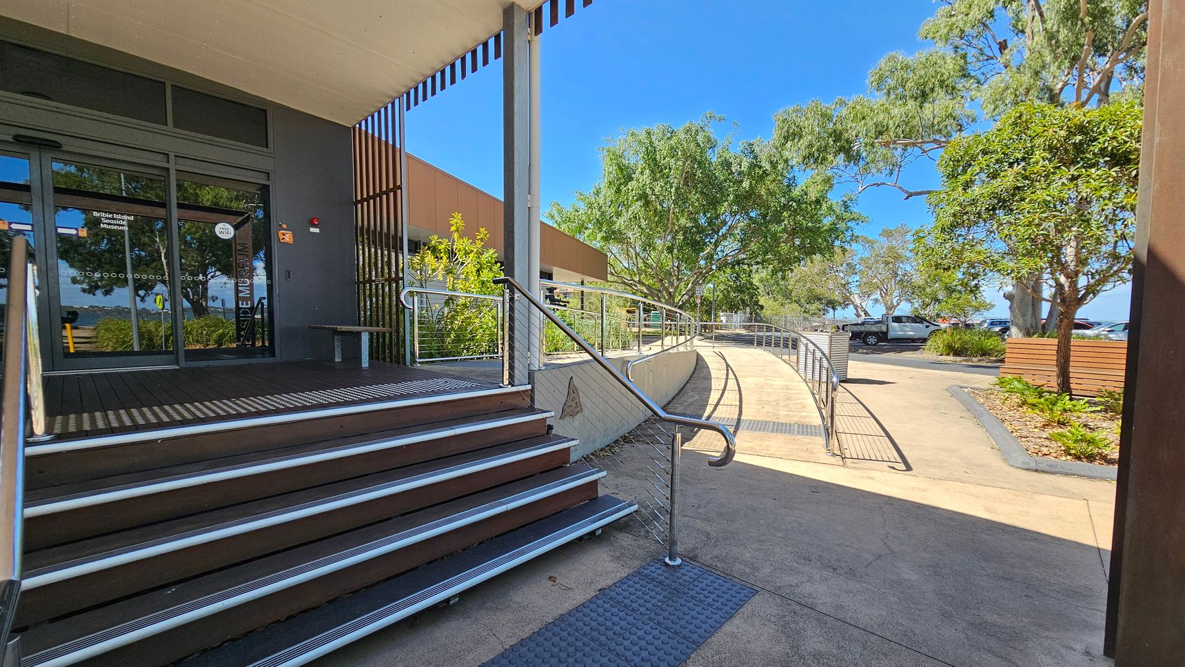 Entrance to a building with a glass door on the left, featuring a sign. A set of stairs with metal railings leads up to the door. To the right, a curved ramp with railings provides an accessible path. The area is surrounded by trees and shrubs, with a parked vehicle visible in the background. The ground is paved, and there is a wooden bench near the greenery. The ceiling above the entrance is flat and extends outward.