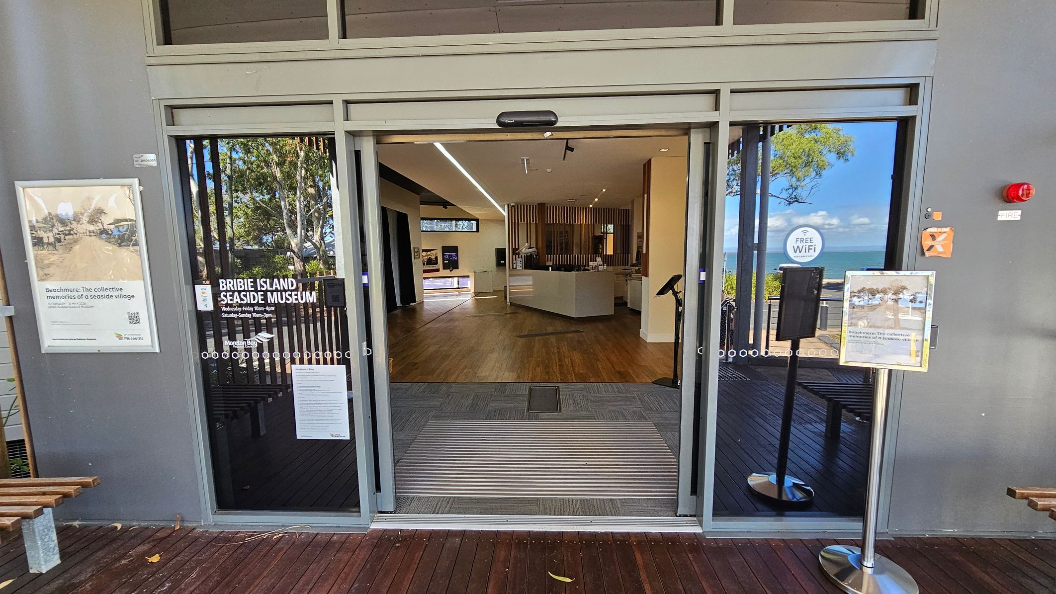 Glass entrance doors to the Bribie Island Seaside Museum, with a poster on the left wall and a sign on a stand to the right. The floor outside is wooden decking, transitioning to a grey mat inside. The interior features wooden flooring, a reception desk, and a display area with modern lighting. A window on the right offers a view of trees and the sea. A "Free WiFi" sticker is visible on the door.