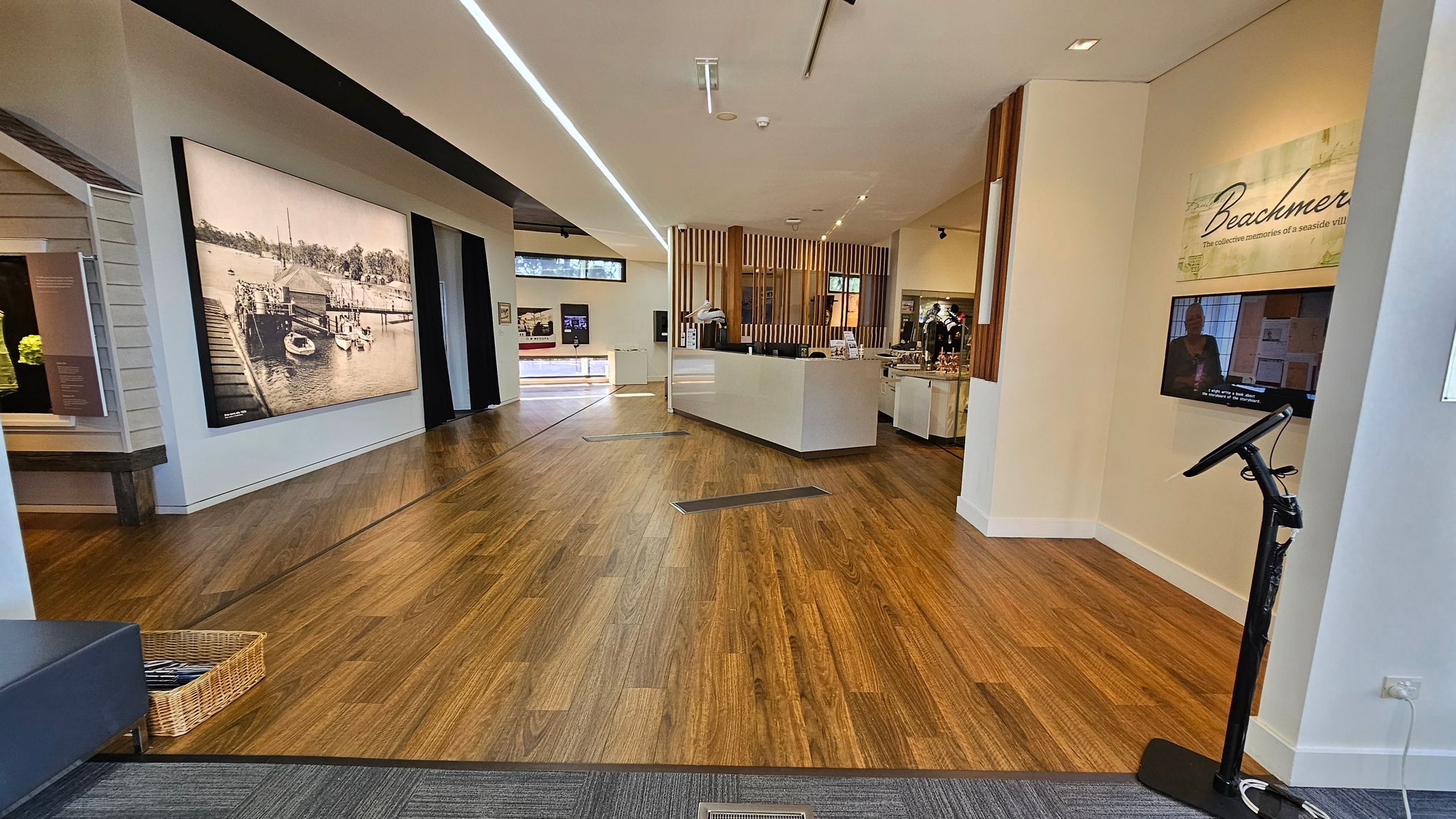 A modern interior with wooden flooring and a white ceiling. On the left, a large black-and-white photograph of a dock scene is displayed on the wall. In the centre, a white reception desk is positioned in front of vertical wooden slats. To the right, a wall-mounted screen displays a video beneath the word "Beachmere." A stand with a touchscreen is nearby. The space is well-lit with overhead lighting, and a basket is placed on the floor near a bench.
