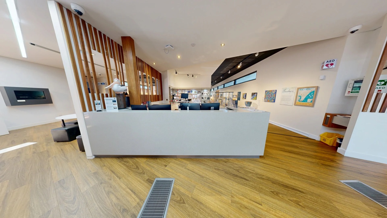 A modern interior with a wooden floor and a white ceiling. In the foreground, a large white reception desk with computer monitors and informational signs. To the left, a wall-mounted screen and some seating. The background features wooden slats and a well-lit area with more seating and displays. On the right, framed pictures and emergency equipment are mounted on the wall. The space is open and well-lit with overhead lights.