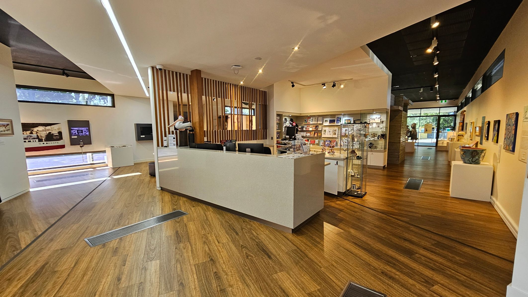 A modern interior with wooden flooring and a high ceiling. On the left, there are display panels and a long window near the ceiling. In the centre, a reception desk with computers and a pelican sculpture. To the right, shelves and glass cases filled with books and items. The background features more displays and a glass entrance with a view of greenery outside. The lighting is a mix of spotlights and natural light.