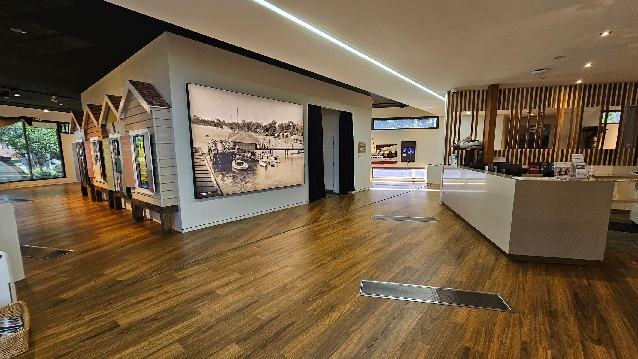 Interior of a modern exhibition space with wooden flooring. On the left, there are small house-like structures with displays and a large historical photograph of a dock scene. The right side features a sleek, white reception desk with informational materials. Behind the desk, vertical wooden slats create a partition. Large windows in the background allow natural light to enter, and the ceiling has recessed lighting.