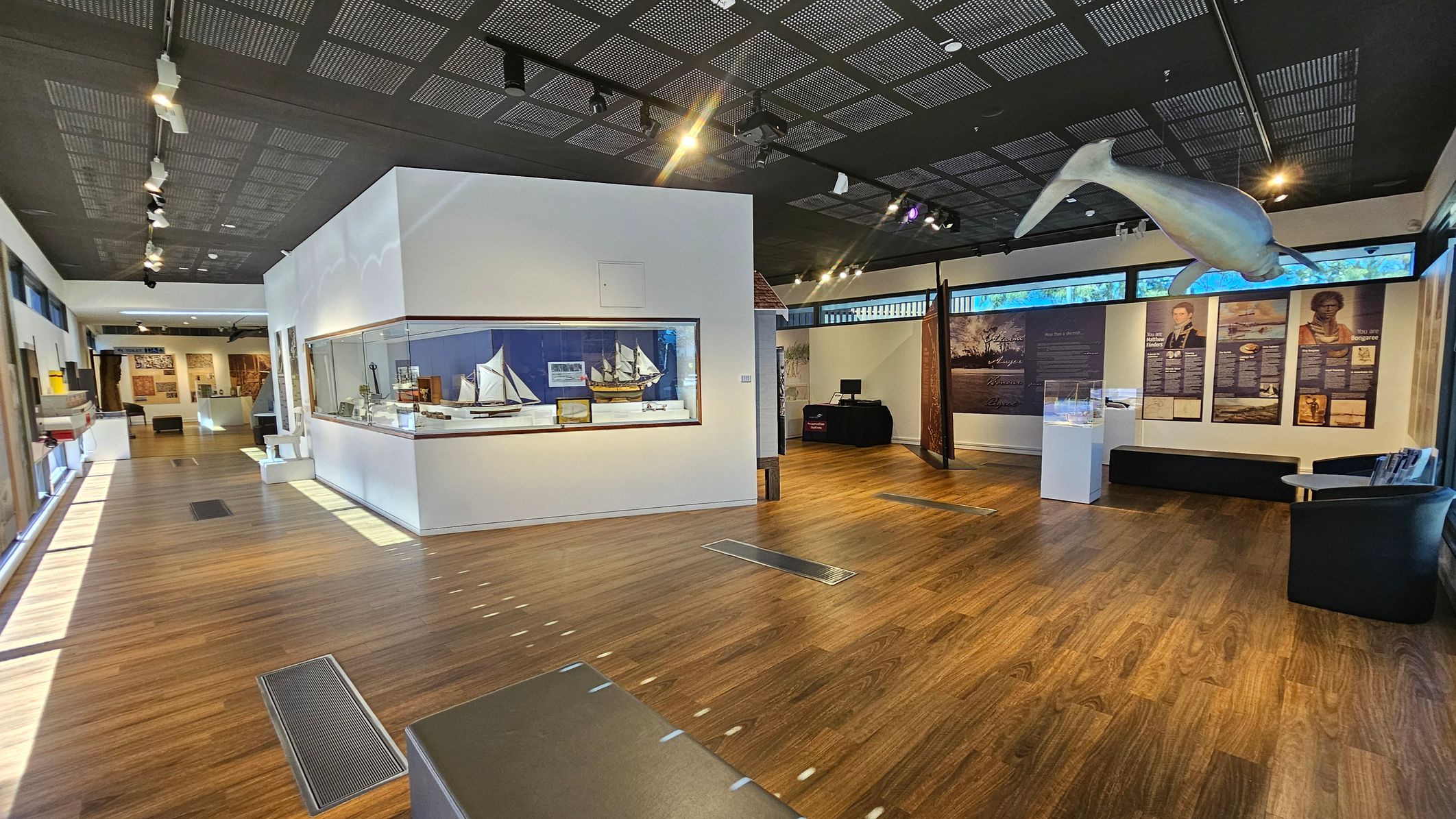 A museum interior with wooden flooring and a dark, grid-patterned ceiling. On the left, a large display case contains model ships. The right side features informational panels and a suspended model of a marine animal. In the background, more exhibits and a small seating area are visible. The space is well-lit with track lighting, and there are windows along the upper walls.