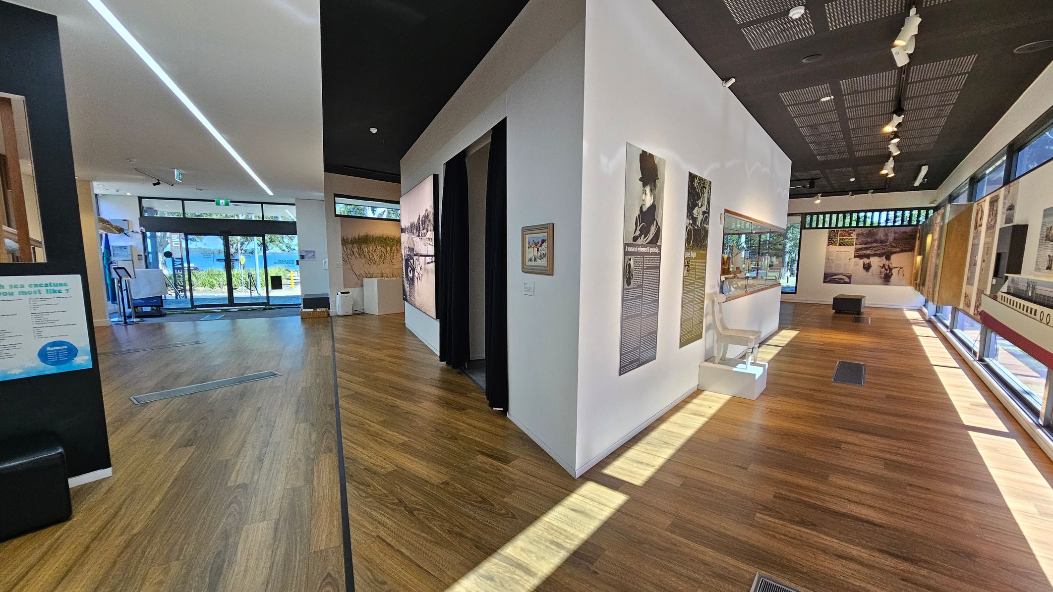 A museum interior with wooden flooring and a black ceiling. On the left, a glass entrance door with a view of trees and water outside. In the centre, a white wall with informational panels and a small framed picture. To the right, more display panels and a white chair on a pedestal. Large windows line the right side, allowing natural light to illuminate the space. Spotlights are mounted on the ceiling, and a black bench is visible in the background.
