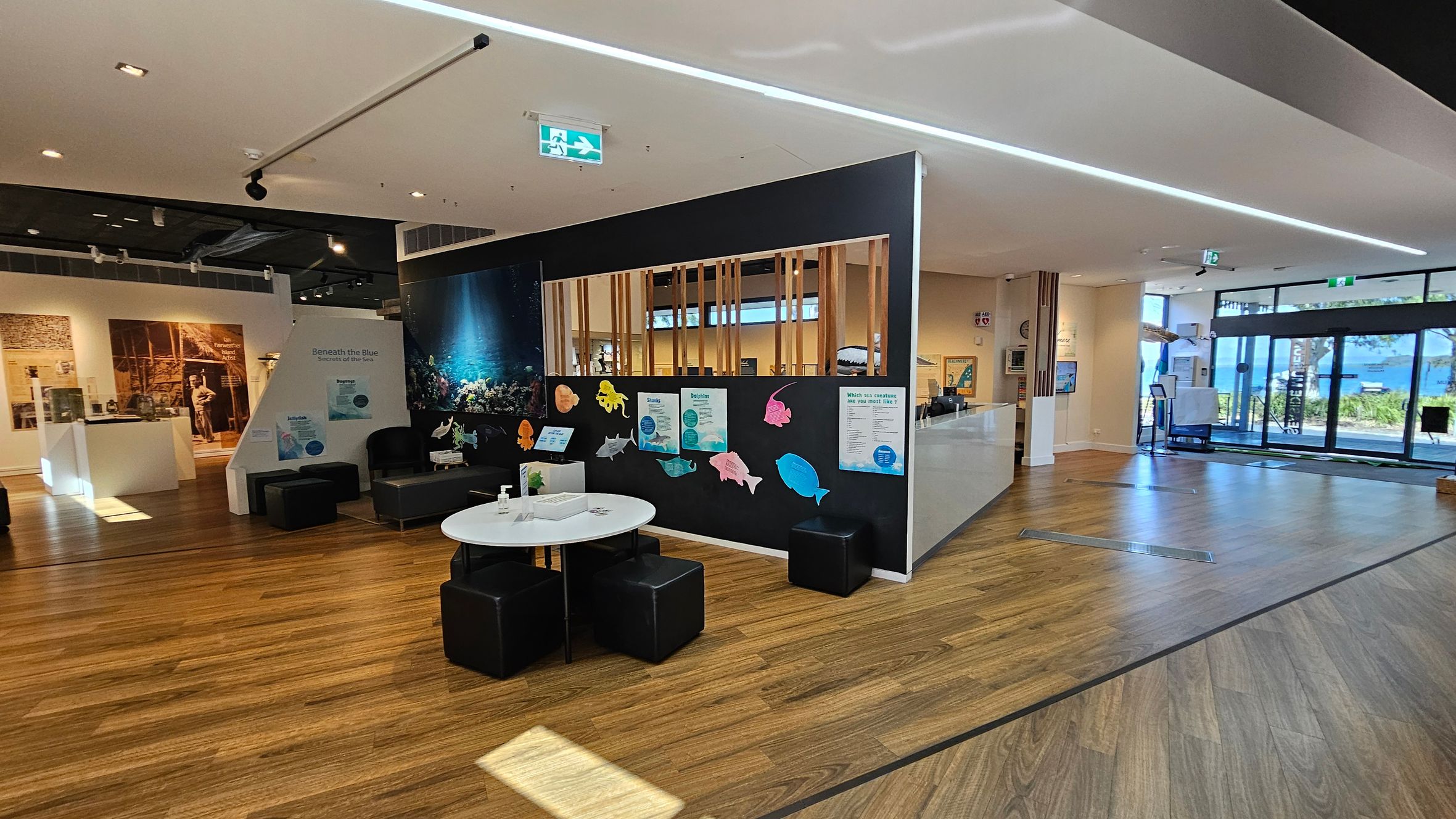 A spacious interior with wooden flooring and a white ceiling. On the left, there is a display area with historical images and artefacts. In the centre, a black partition wall features colourful fish cut-outs and informational posters. A round white table with black cube seats is in front. On the right, large glass doors lead outside, allowing natural light to illuminate the space. The ceiling has recessed lighting and an emergency exit sign.