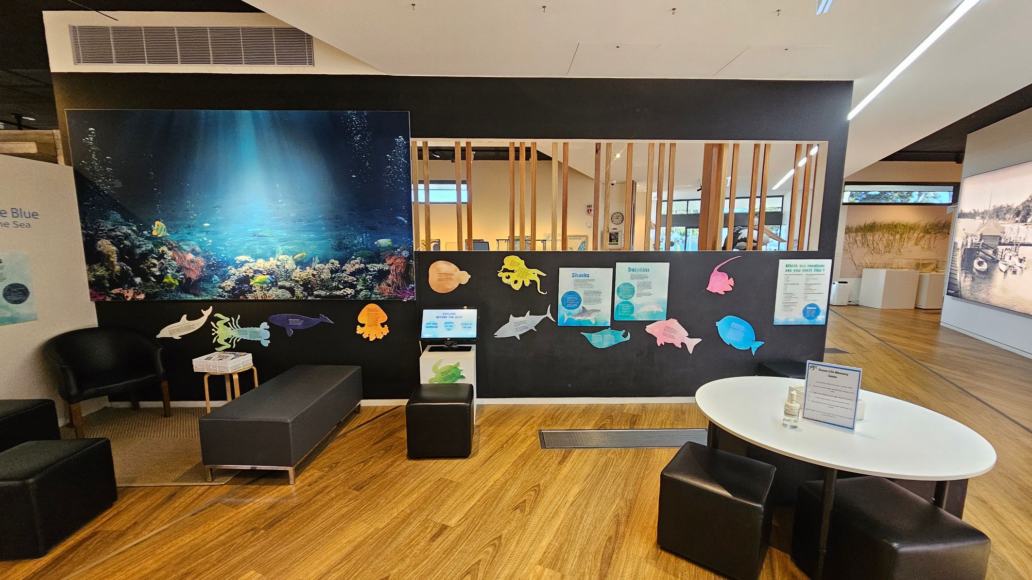 On the left, there's a seating area with black chairs and a small table. A large underwater-themed mural with coral and fish is on the wall. The middle section features a black partition with colourful cutouts of sea creatures and informational posters. A small screen is mounted on the partition. On the right, a white oval table with black stools is placed on a wooden floor. The background shows a room with wooden slats and large windows.