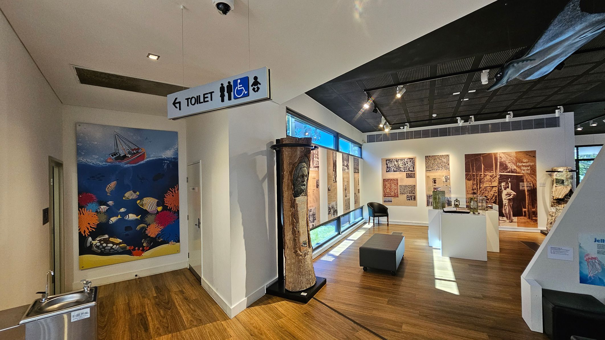 A museum interior with wooden flooring. On the left, a colourful mural of an underwater scene with fish and coral. Above, a sign indicates the direction to the toilet, with symbols for accessibility and baby changing. In the centre, a carved wooden sculpture stands against a wall. The right side features large windows and displays with text and images, including a large photograph of a person. A grey bench and a black chair are placed near the displays. The ceiling is dark with spotlights.