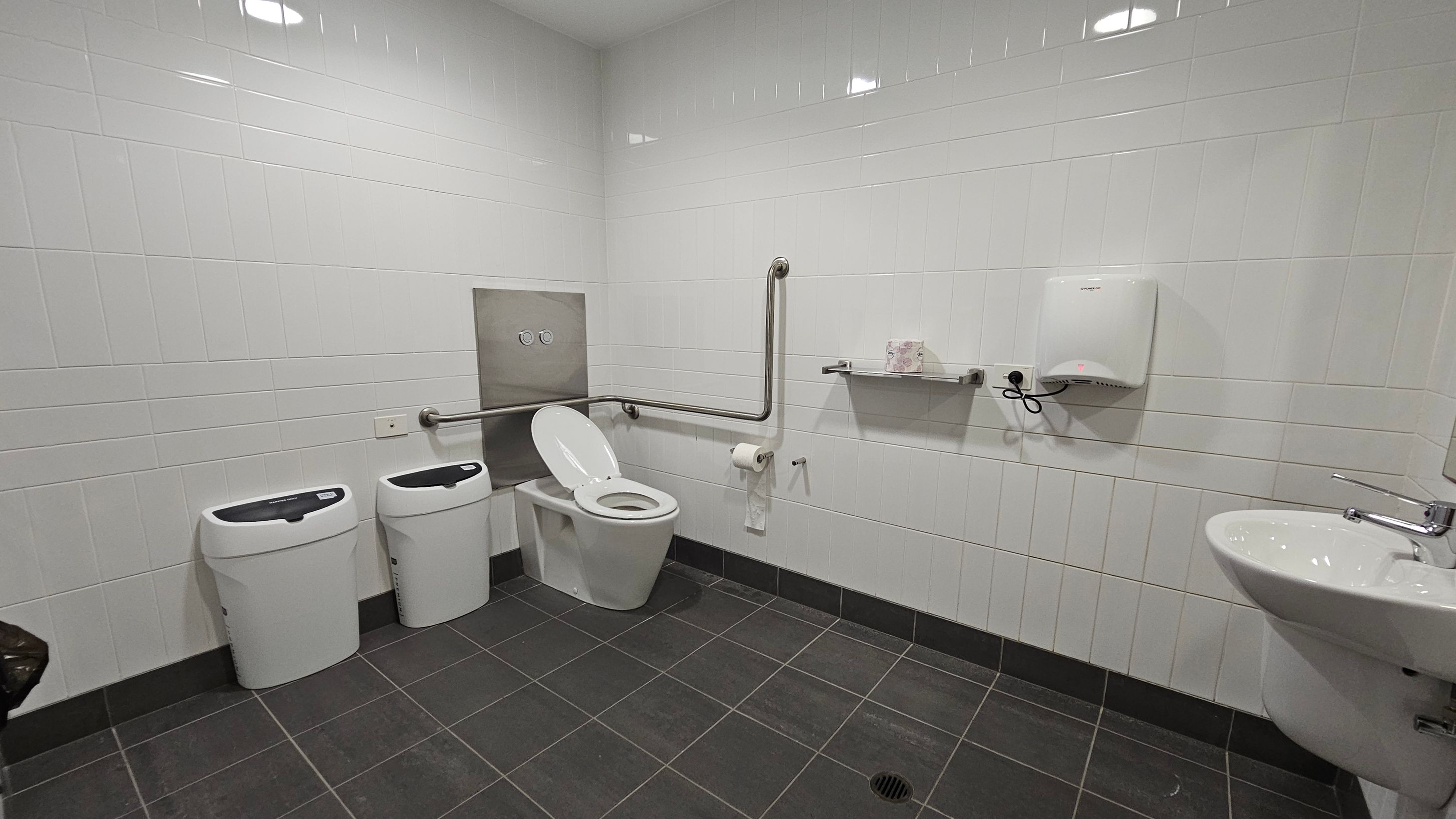 A public restroom with white tiled walls and a dark tiled floor. On the left, there are two white bins. In the centre, a toilet with a raised seat and a grab bar beside it. Above the toilet, a flush panel is visible. To the right, a shelf holds a box of tissues, and a wall-mounted hand dryer is installed. Further right, a white sink with a chrome tap is attached to the wall. The ceiling has recessed lighting.