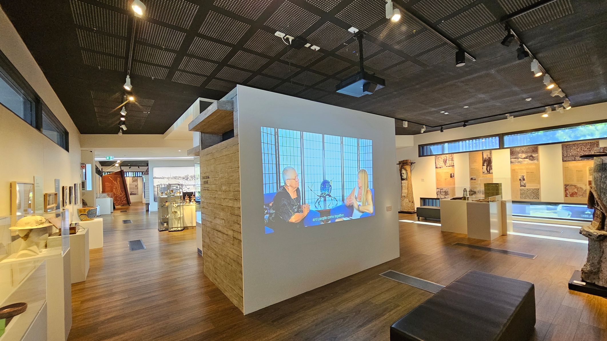 A modern museum interior with wooden flooring and a dark, perforated ceiling with spotlights. In the centre, a white wall displays a video of two people in conversation. To the left, glass cases exhibit various artefacts, while the right side features informational panels and sculptures. The background shows more displays and a large window allowing natural light. A black bench is positioned in the foreground.