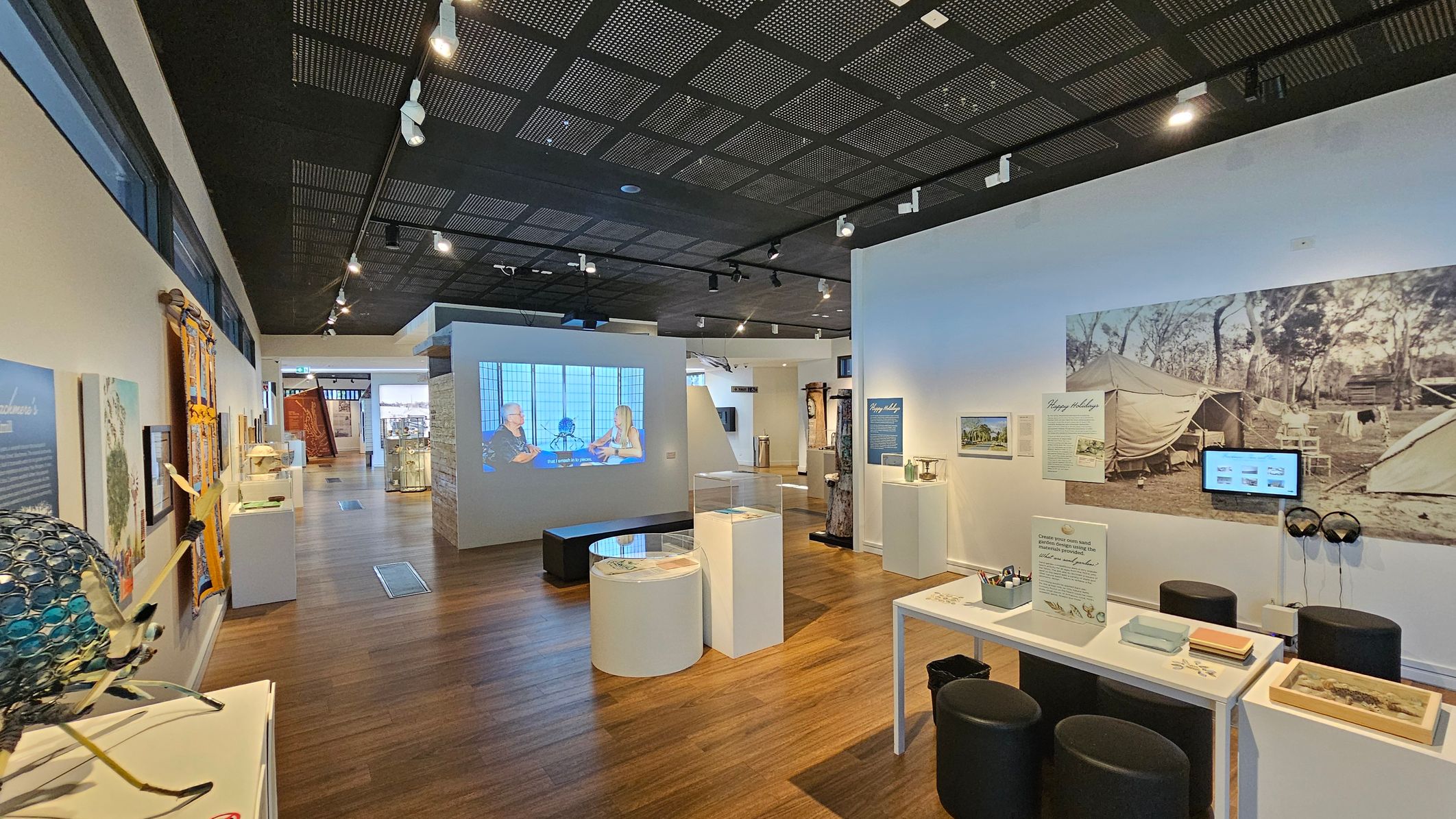 A museum interior with wooden flooring and a black perforated ceiling. On the left, there are various exhibits, including a colourful textile piece and a sculpture made of circular elements. In the centre, a video is projected onto a wall, with seating nearby. On the right, a large photograph of a campsite is displayed, accompanied by informational panels and a small screen with headphones. Several tables and stools are arranged for interactive activities.