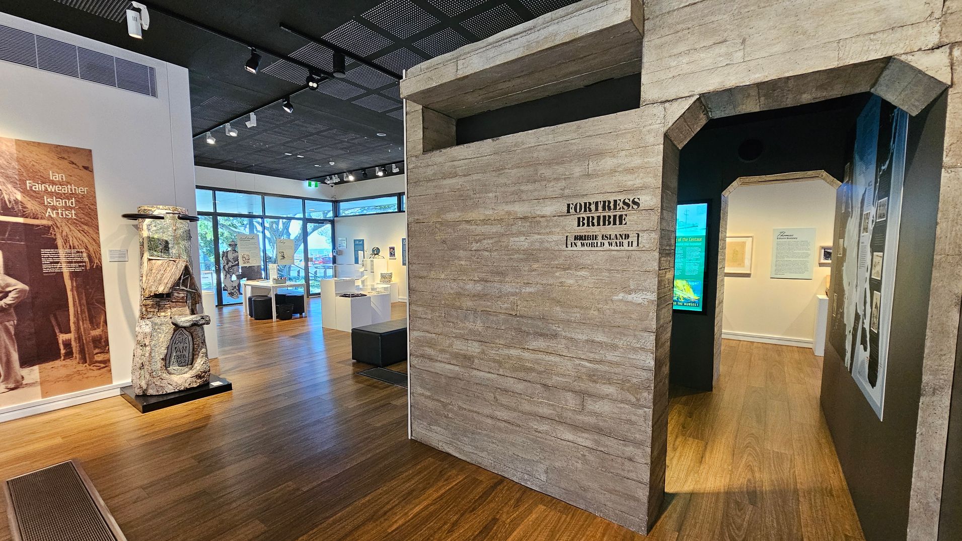 A museum interior with wooden flooring and a dark ceiling with spotlights. On the left, a display features a large poster about Ian Fairweather, an island artist, alongside a sculptural piece. In the centre, a structure resembling a bunker entrance is labelled "Fortress Bribie, Bribie Island in World War II." The right side shows an open doorway leading to an exhibit area with framed pictures and informational panels. Large windows in the background allow natural light in.