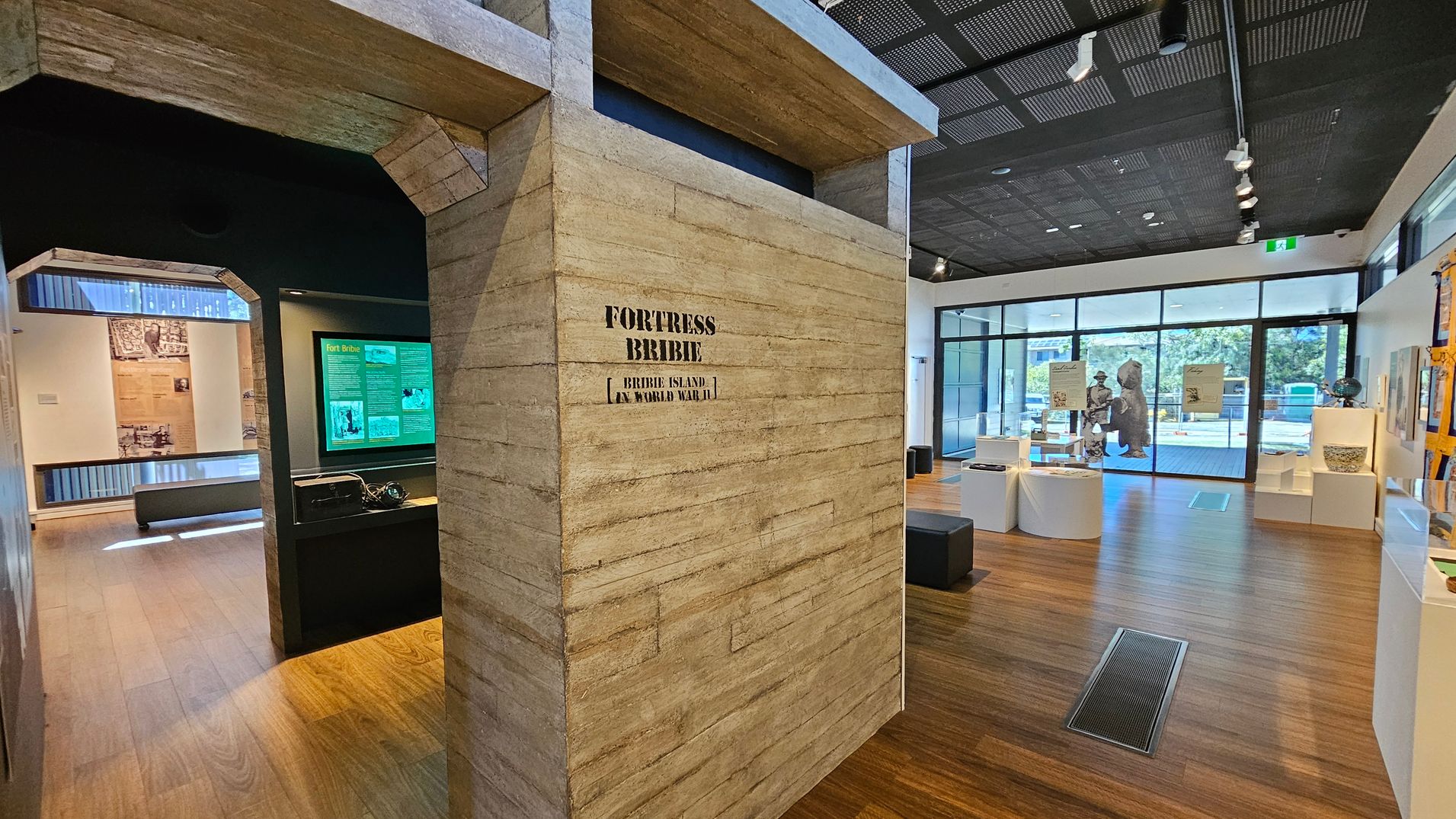 A museum interior with a wooden floor and a dark, perforated ceiling. In the centre, a large structure with "FORTRESS BRIBIE" written on it. To the left, an exhibit with green informational panels and historical images. On the right, a spacious area with display cases and a large window showing outdoor greenery. The room is well-lit with track lighting, and there are various artefacts and informational boards throughout.