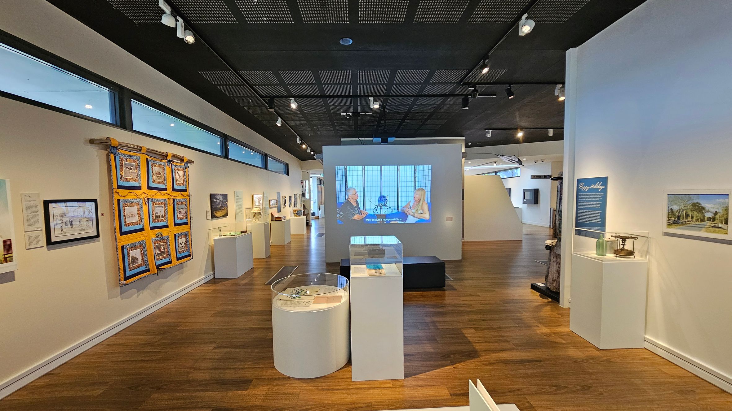 A gallery with wooden flooring and a black perforated ceiling. On the left, a colourful quilt hangs on the wall alongside framed artworks. In the centre, a video is projected onto a wall, showing two people in conversation. Below the projection, there are display cases on white pedestals. On the right, a display case with objects and a blue information panel is visible, along with a framed landscape picture. The space is well-lit with track lighting.
