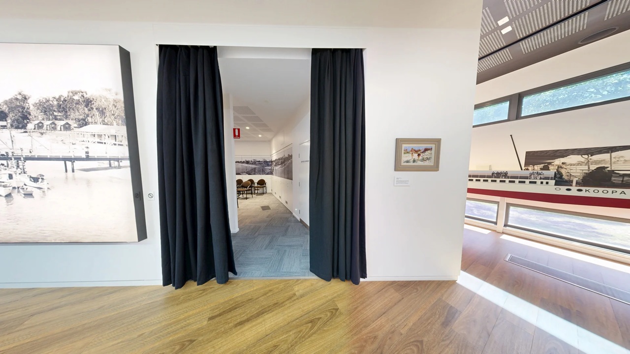 A room with wooden flooring and white walls. On the left, a large black-and-white photograph of a waterfront scene is displayed. In the centre, a doorway with black curtains leads to another room with blue carpet and chairs. On the right, a small framed painting is mounted on the wall, and a large mural of a ship with the name "KOOPA" is visible. The ceiling has a modern design with recessed lighting.