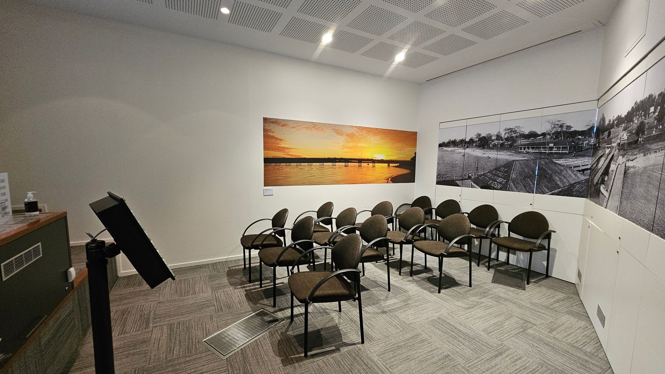 A small room with a series of brown chairs arranged in rows on a carpeted floor. On the left, there's a podium with a tilted stand. The back wall features a large photograph of a sunset over a pier. The right wall displays a black-and-white panoramic image of a waterfront scene. The ceiling has recessed lighting and a grid pattern.