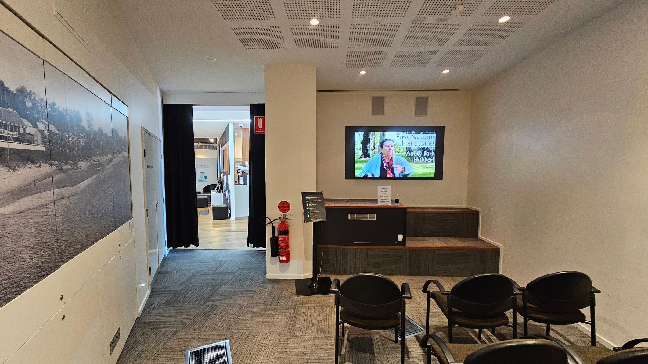 A room with a television mounted on the right wall displaying a video titled "First Nations Elder Stories." Below the TV is a wooden bench with a fire extinguisher and a small sign. Four black chairs are arranged in front of the bench. On the left wall, there is a large black-and-white photograph of a beach scene. The floor is carpeted with a patterned design, and the ceiling has recessed lighting. A hallway leads to another area with wooden flooring.