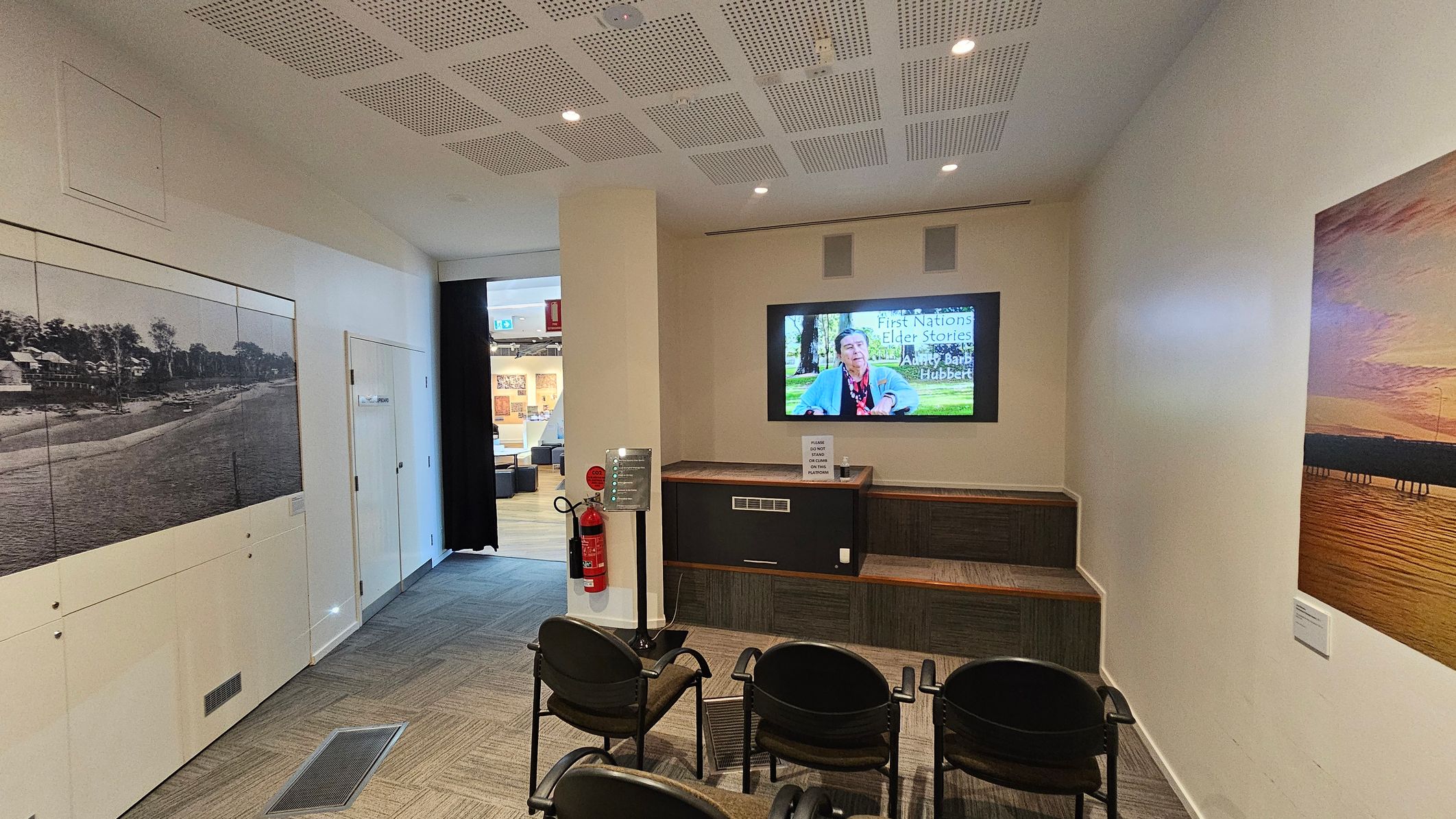 A small room with a screen on the right wall displaying a video titled "First Nations Elder Stories." Below the screen is a wooden bench. Four black chairs are arranged in front of the screen. On the left wall, there is a large black-and-white photograph of a beach scene. A fire extinguisher is mounted near the entrance. The ceiling has a grid pattern with recessed lights. The floor is carpeted with a geometric pattern. An open doorway leads to another room with more displays.