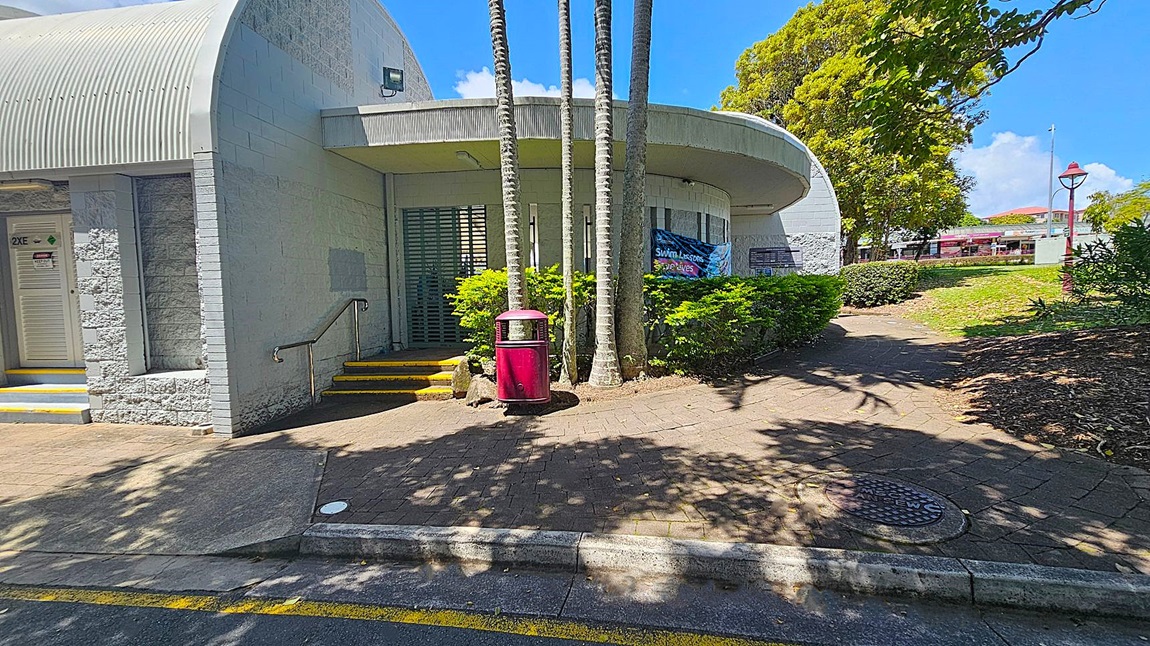 A building with a curved roof and light grey brick walls is on the left. A door marked "2XE" is visible with steps leading up to it. In the centre, several tall palm trees stand in front of a green metal gate, with a red bin nearby. Bushes and a banner advertising swim lessons are in front of the building. On the right, a paved path leads through a grassy area with trees and a red lamp post. Shadows of trees are cast on the ground.